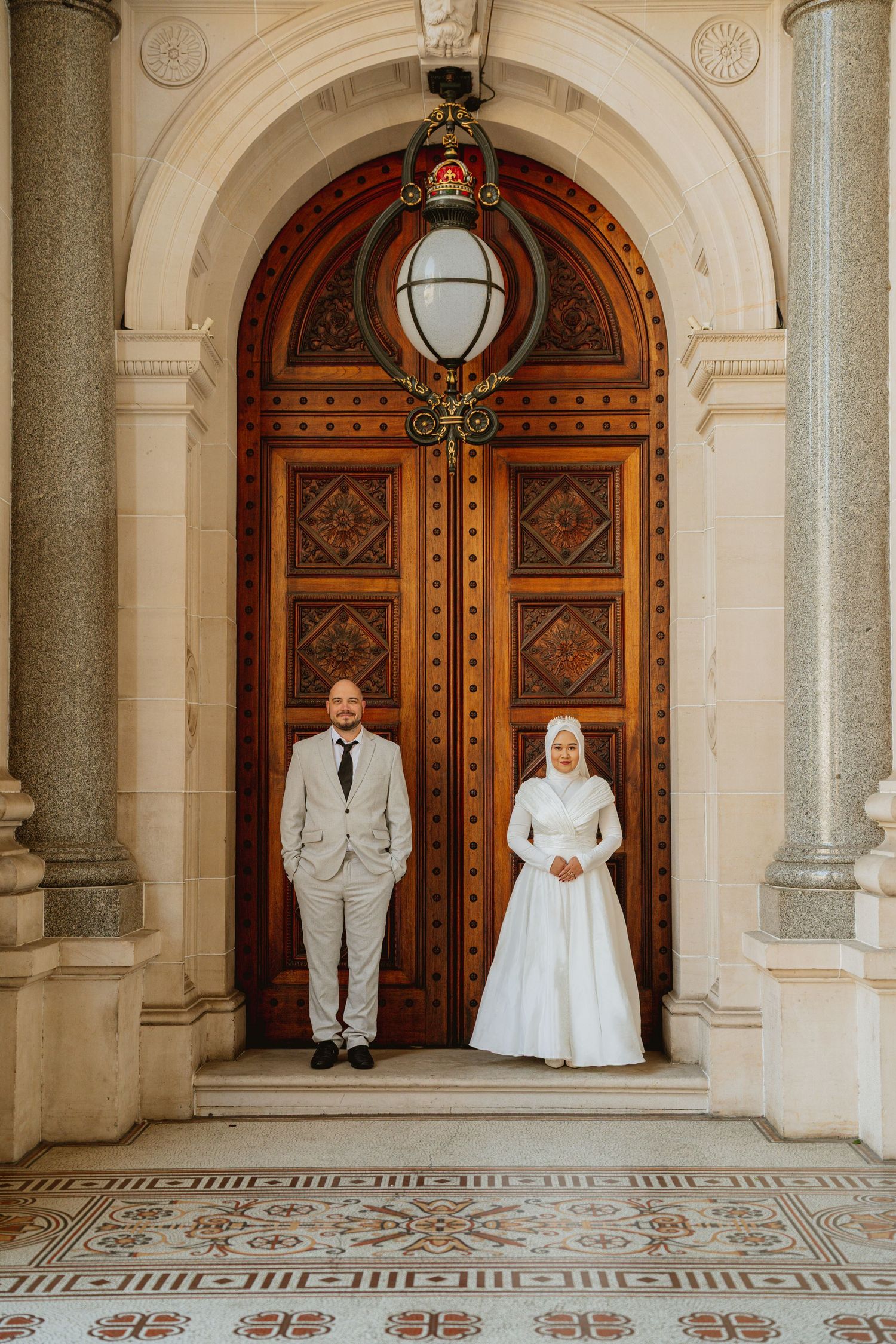 Bride in white hijab gown and groom in grey suit stand before ornate wooden door at elegant wedding venue.
