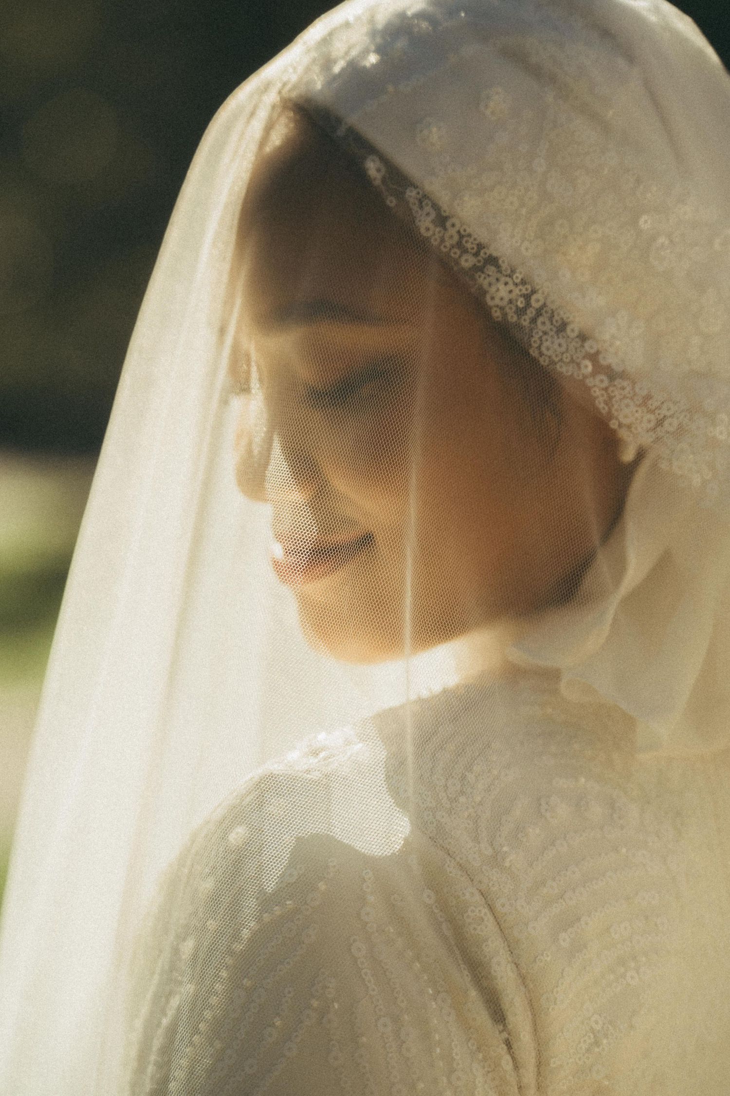 Smiling bride in delicate lace hijab and white gown bathed in soft natural light at outdoor wedding.