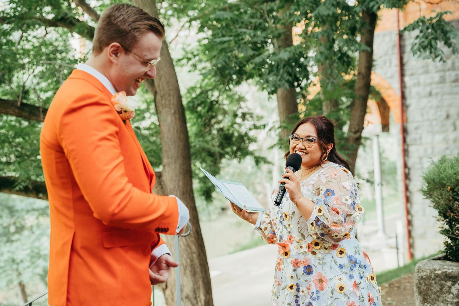 Groom in orange suit listens as woman in floral dress reads vows during outdoor wedding ceremony.