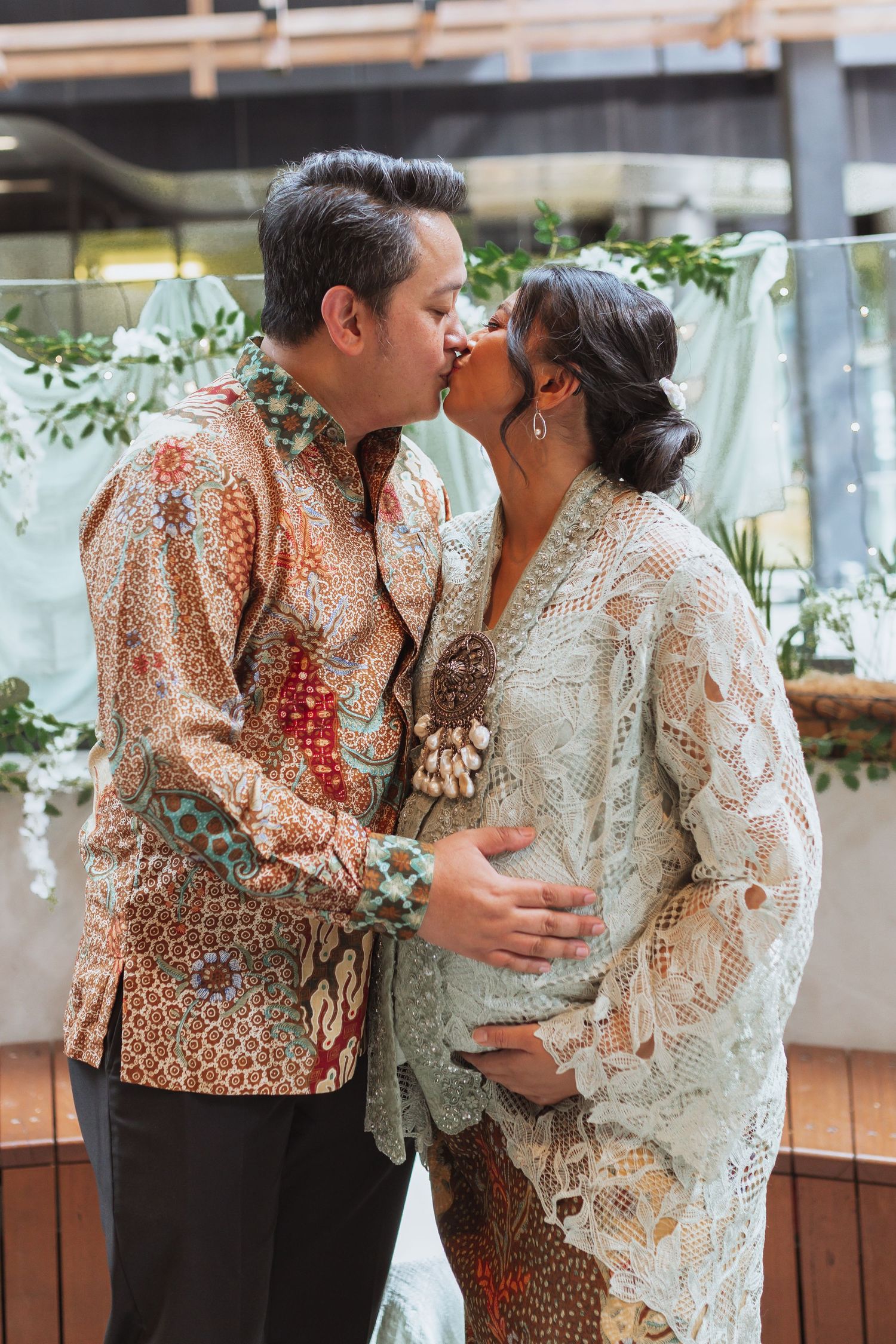 Two people in patterned lace and floral outfits embracing in a bright indoor setting.