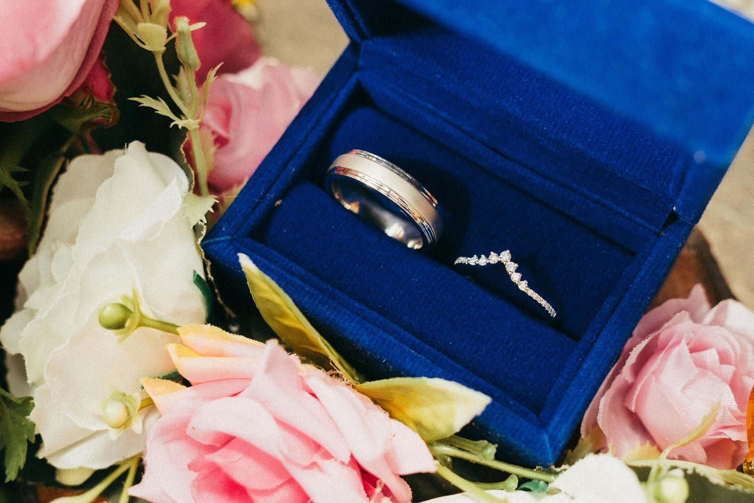 Two wedding rings in a blue velvet box surrounded by pink and white flowers.