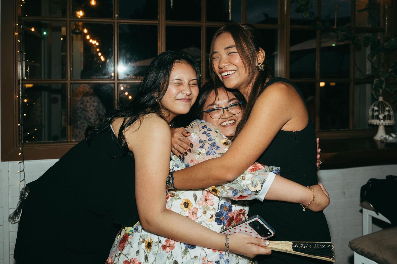 Three young women share a warm group hug at a festive indoor event with string lights.