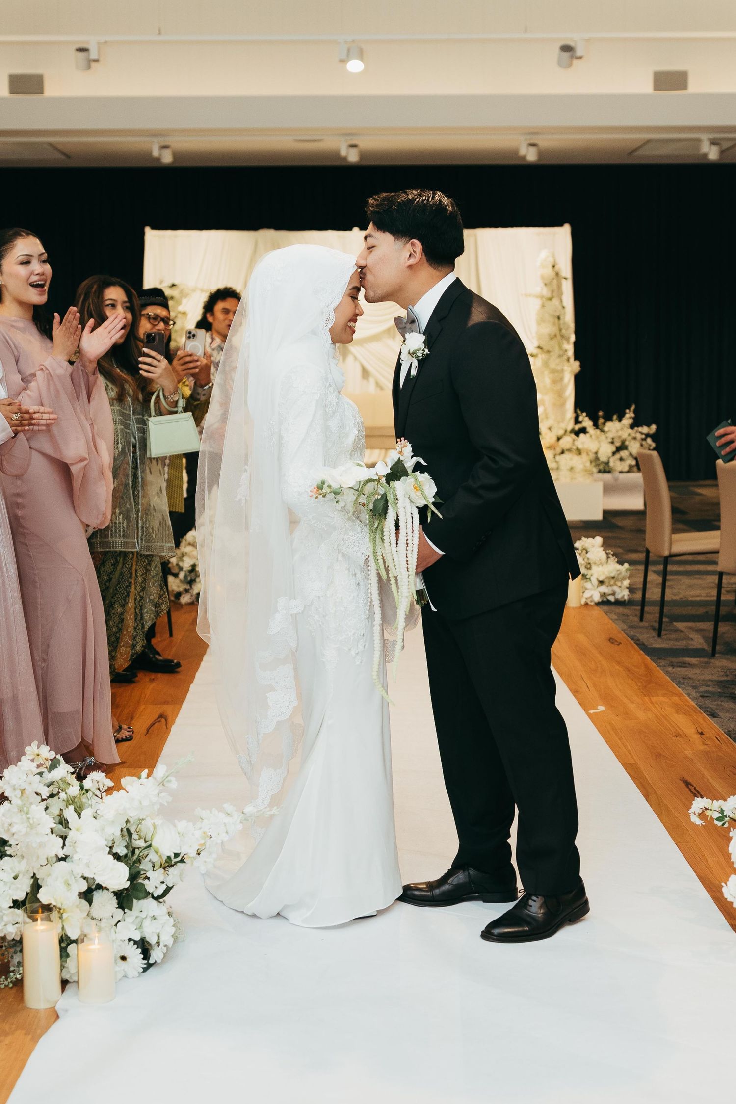 Muslim groom kissing bride on forehead during wedding reception as guests applaud along white floral aisle.