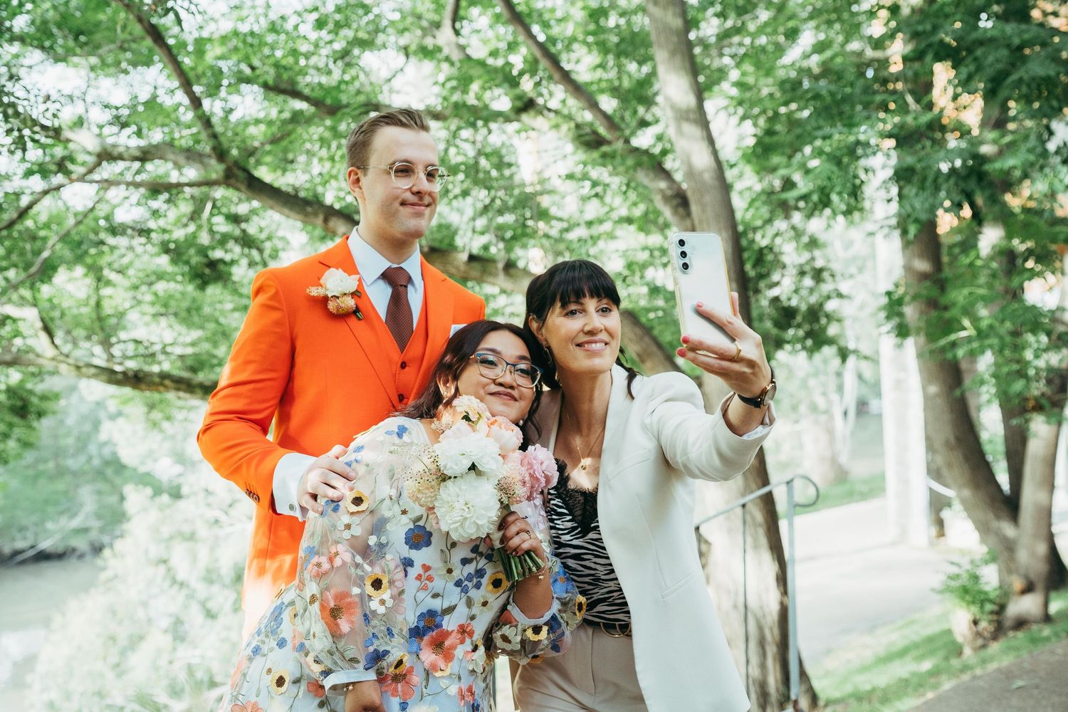 A bride in a floral dress takes a selfie with guests outdoors under green trees.