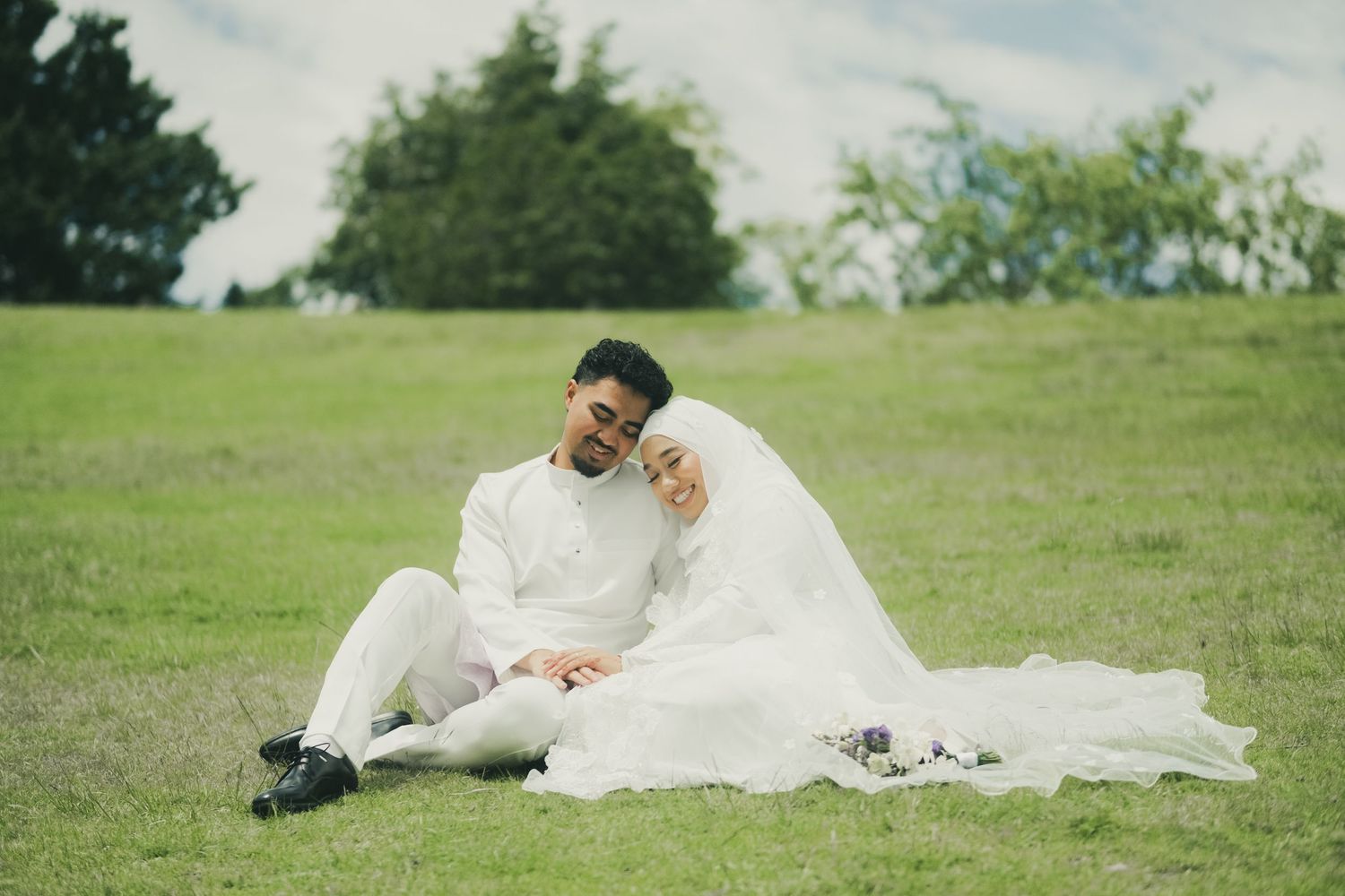 Newlywed Malay couple in matching white outfits sitting together on green grass for wedding photos.