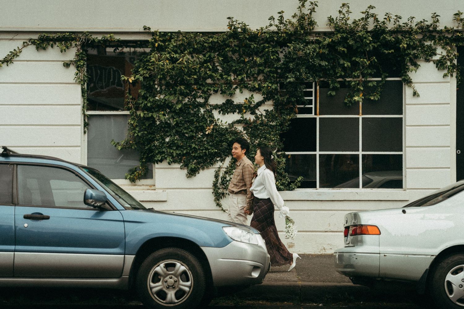 A person walking between parked cars on a city street with ivy-covered white building in the background.