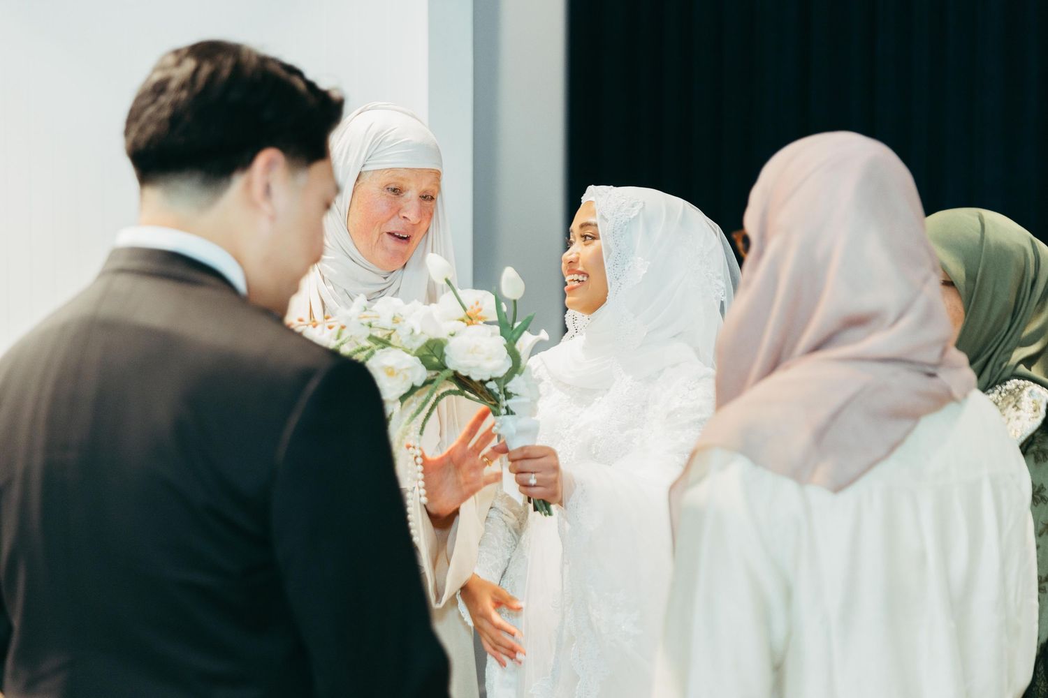 Muslim bride in white hijab gown greeting guests while holding white flower bouquet.