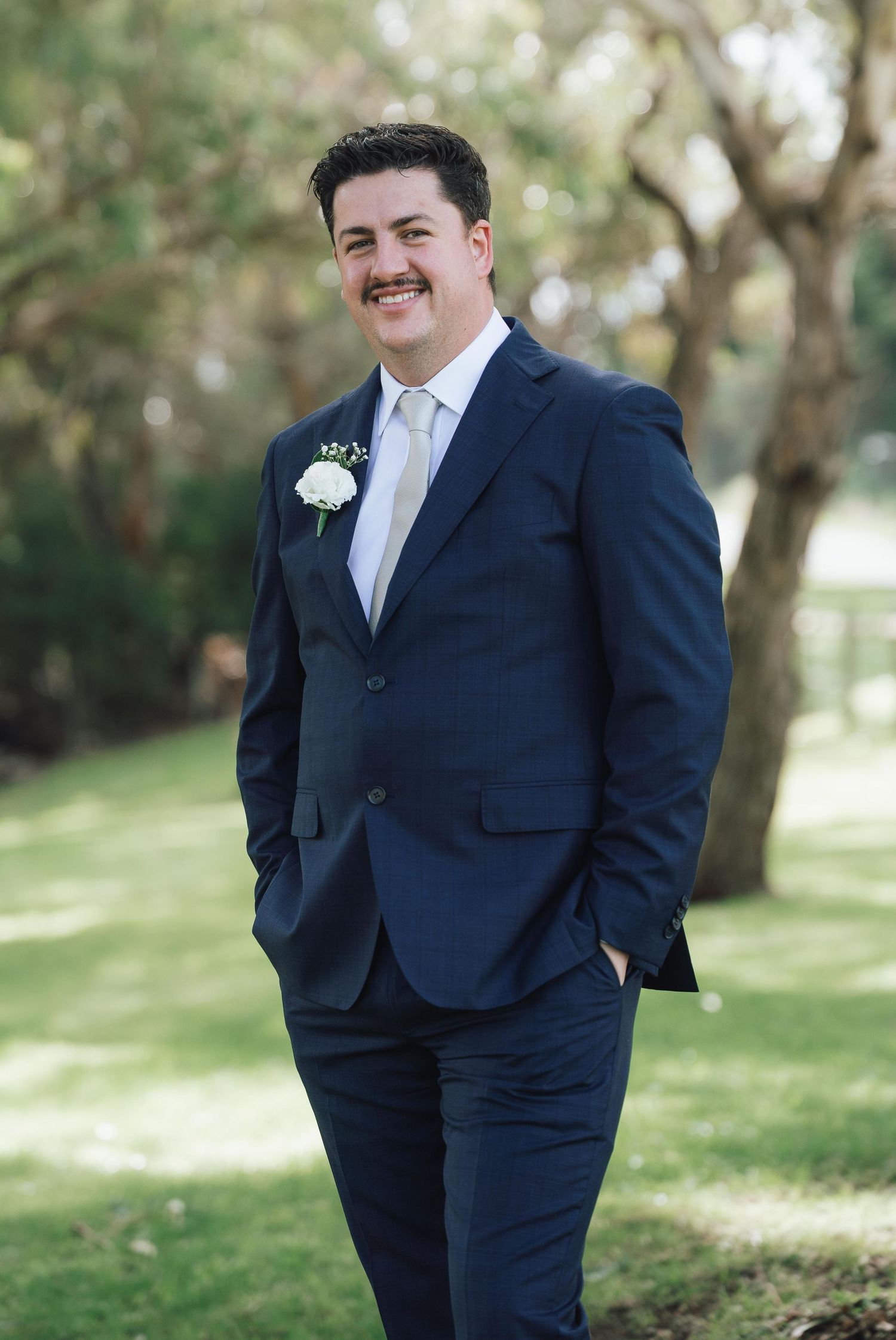 Handsome groom in navy blue suit with white boutonniere smiling outdoors on wedding day.