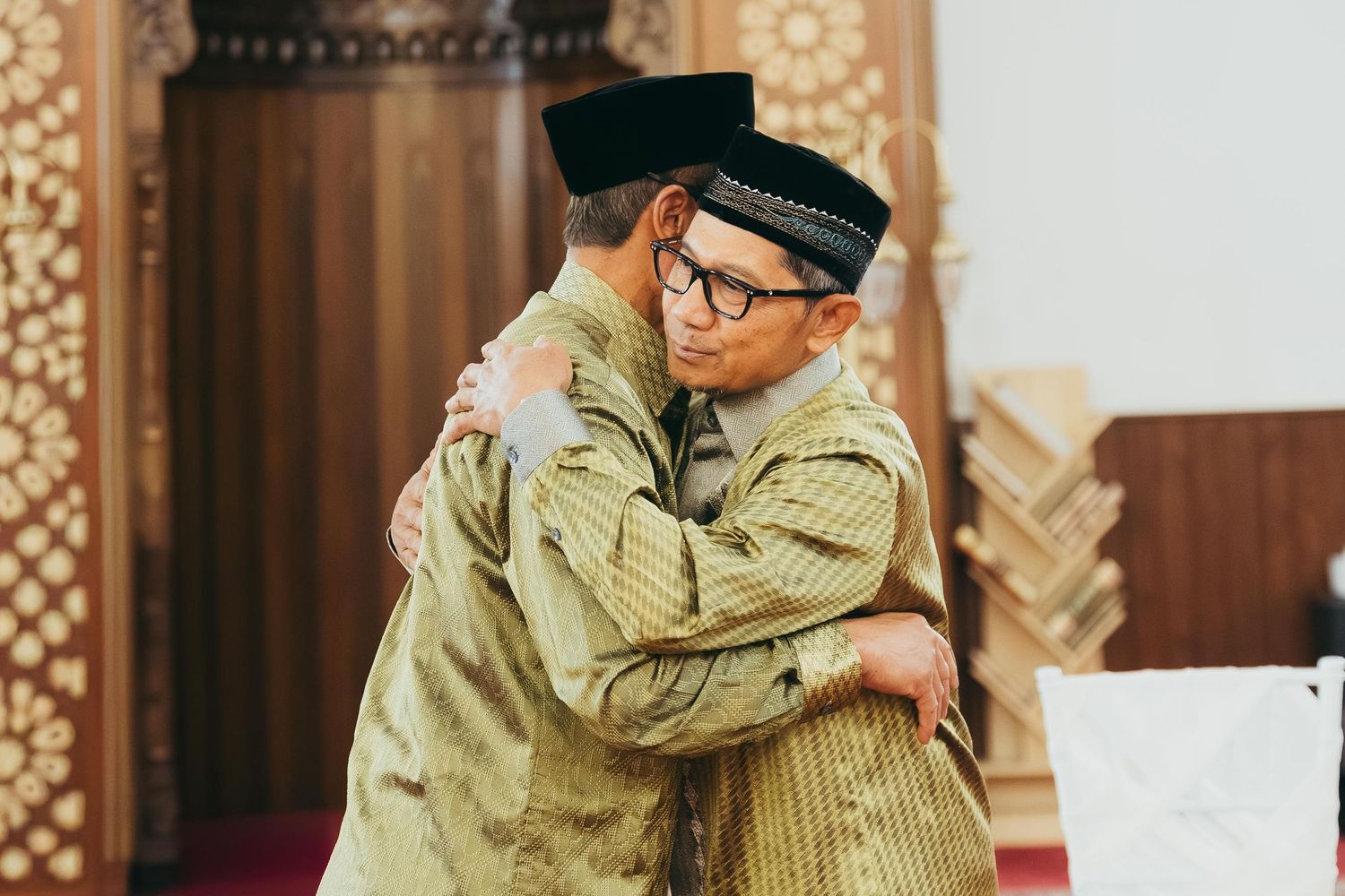 Two men in traditional Islamic attire sharing a warm embrace inside a mosque during Eid celebration.