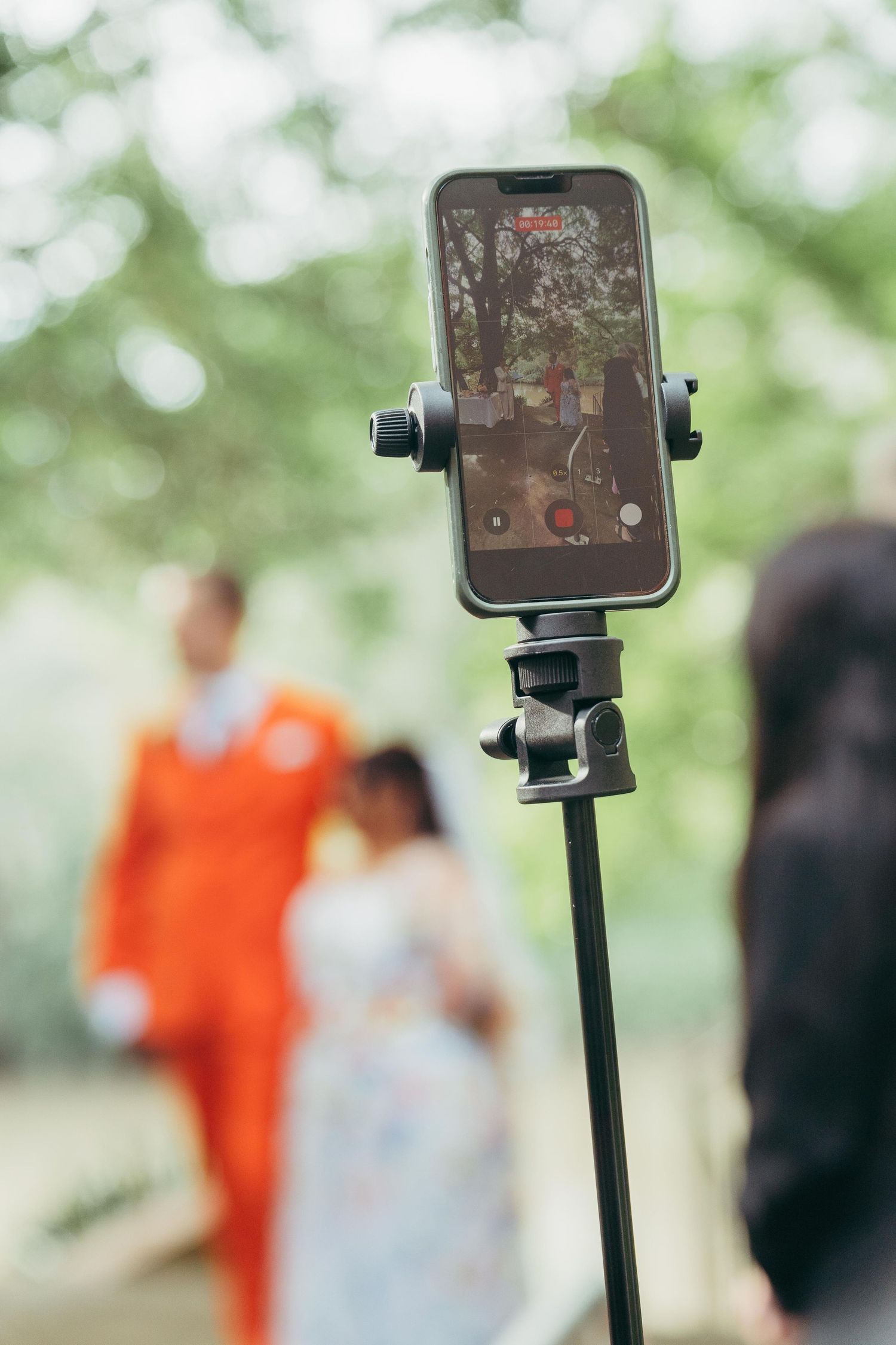 Smartphone on tripod records wedding couple in orange suit and floral dress in lush garden setting.