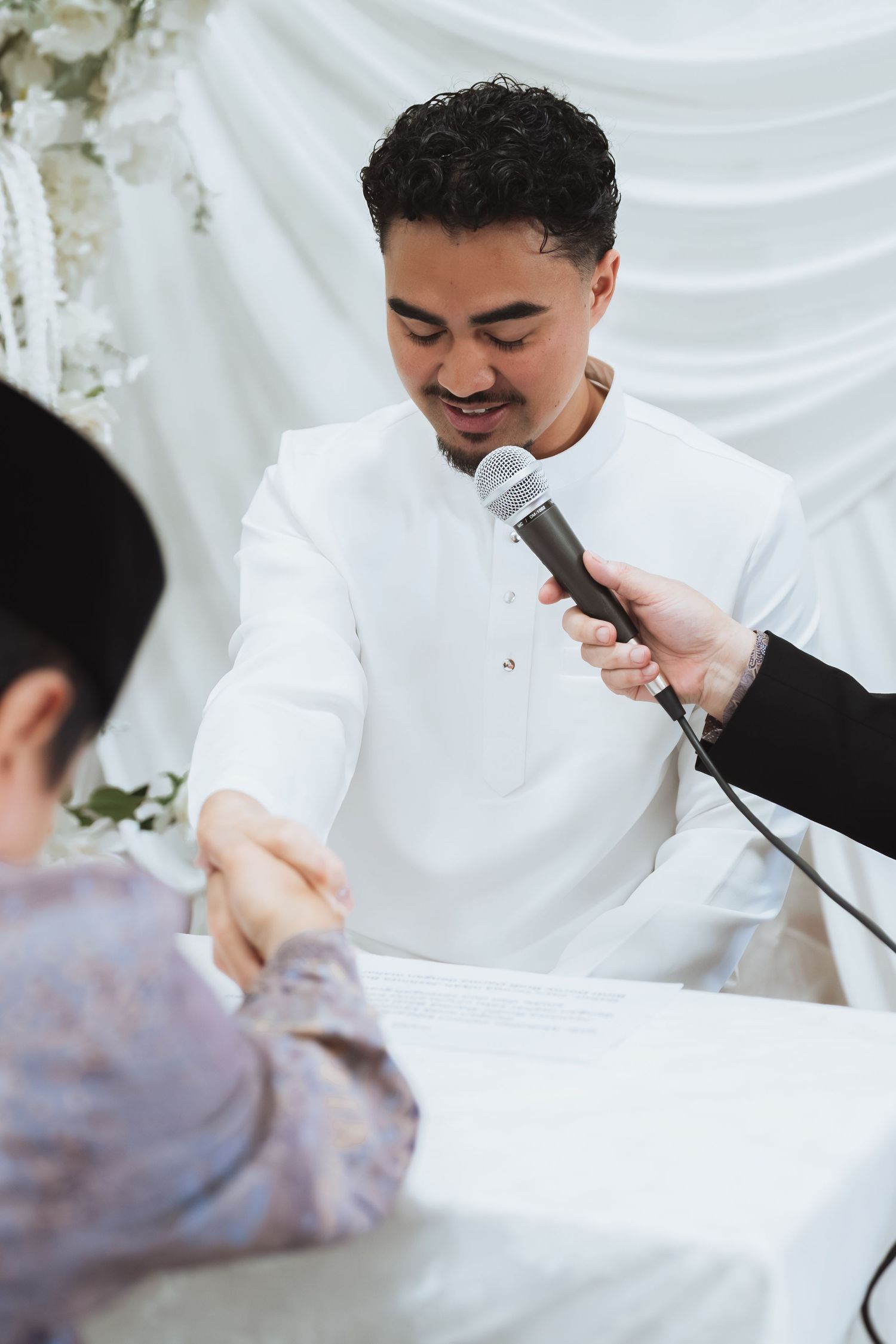 Smiling groom in white baju melayu shaking hands during a traditional Malay wedding nikah ceremony.
