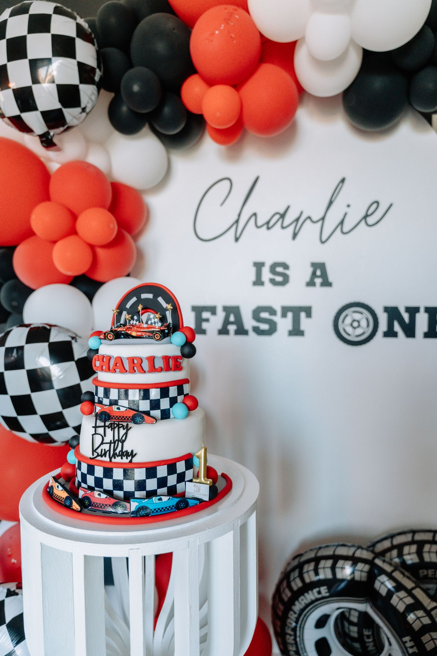 Racing themed birthday party setup with checkered balloons, orange and black balloon garland, and a three-tiered race car cake.