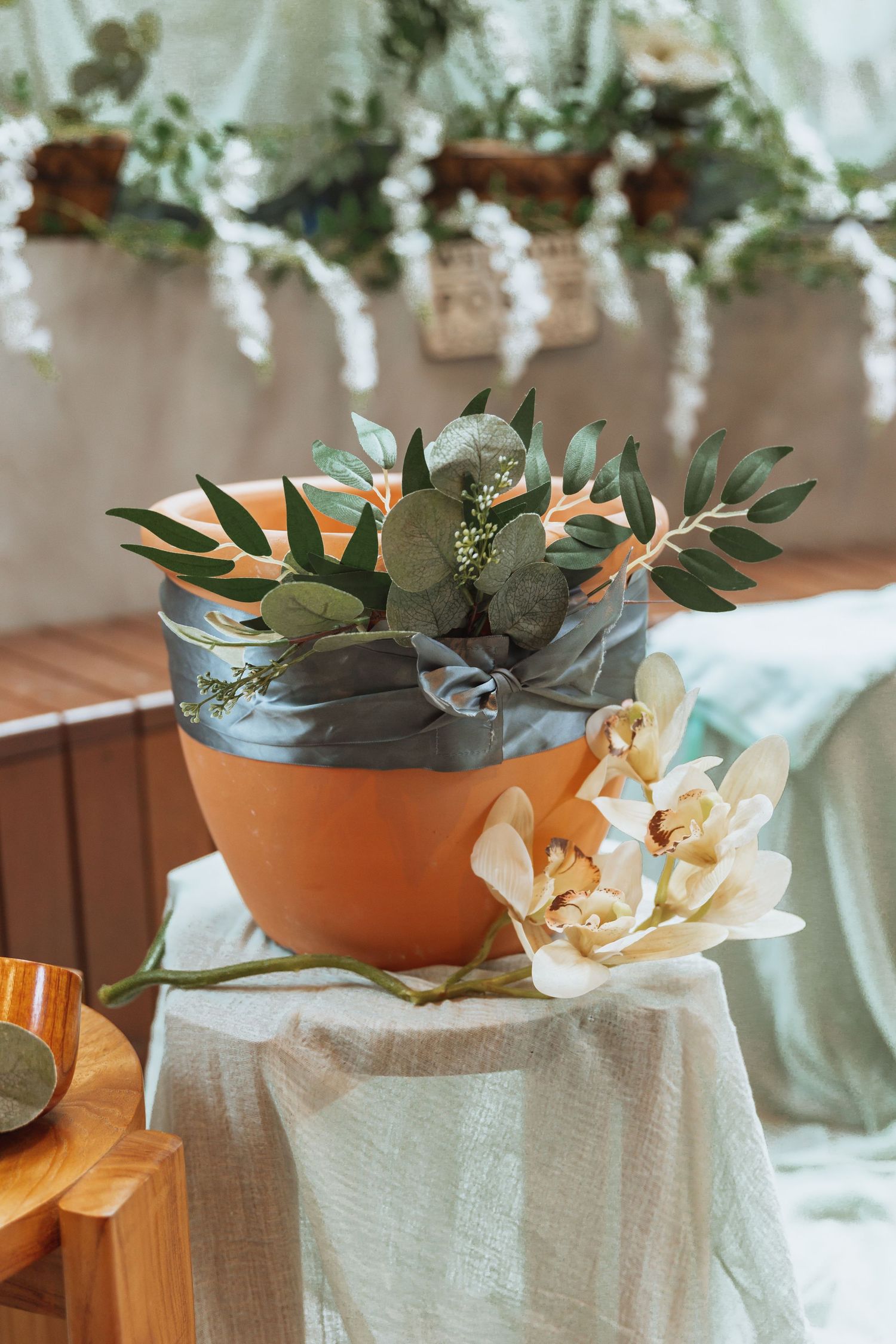 A terracotta pot with eucalyptus leaves and floral arrangements sits on a white cloth against a soft background.
