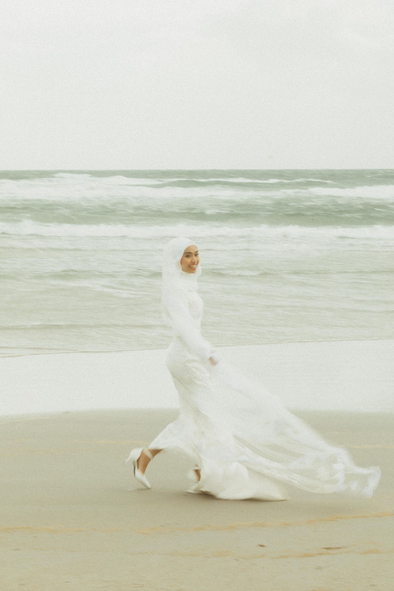 Muslim bride in flowing white wedding gown and hijab walking gracefully along a beach with ocean waves.