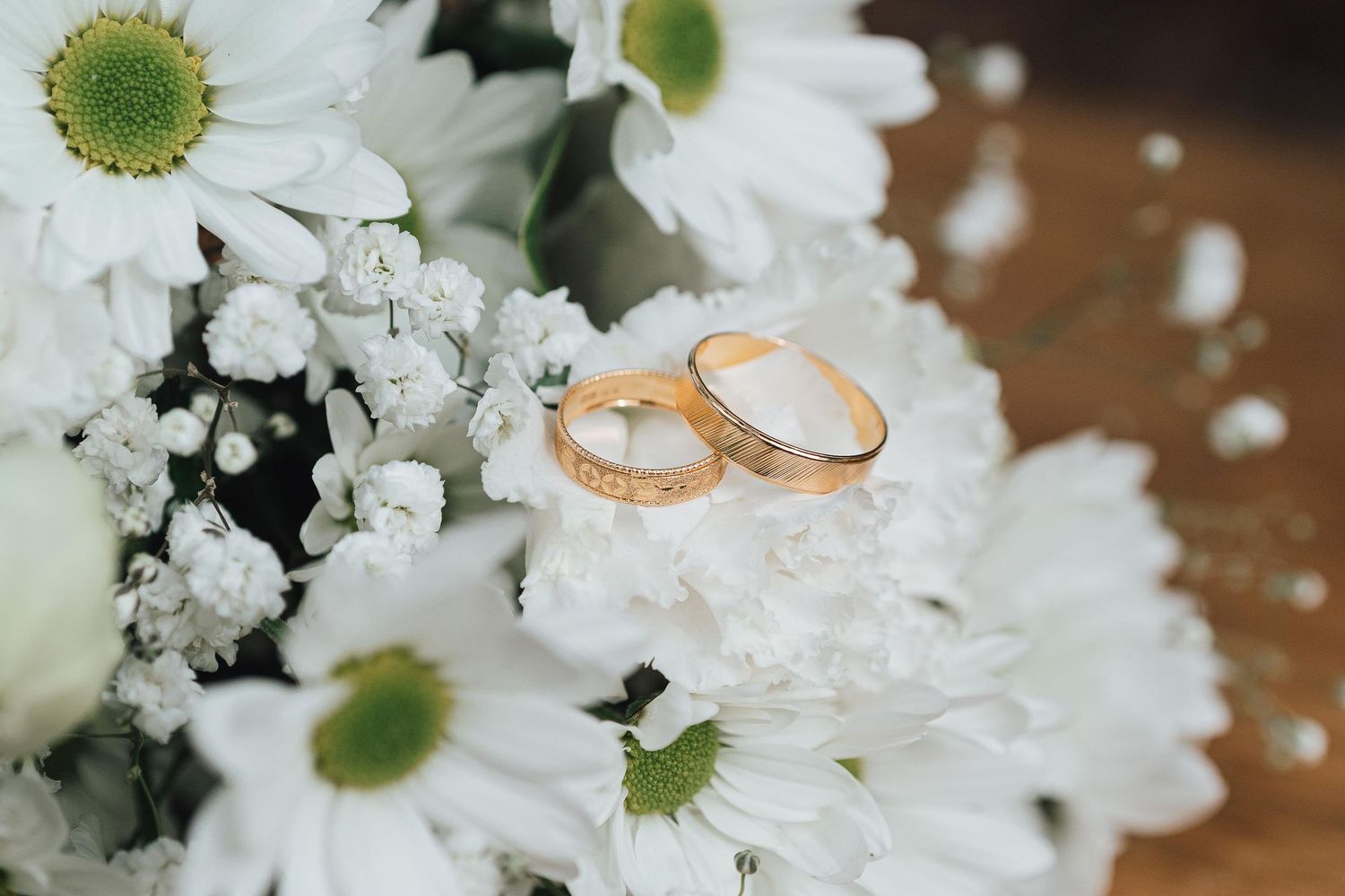 Two gold wedding rings resting on white daisy bouquet, elegant bridal floral arrangement for wedding ceremony.