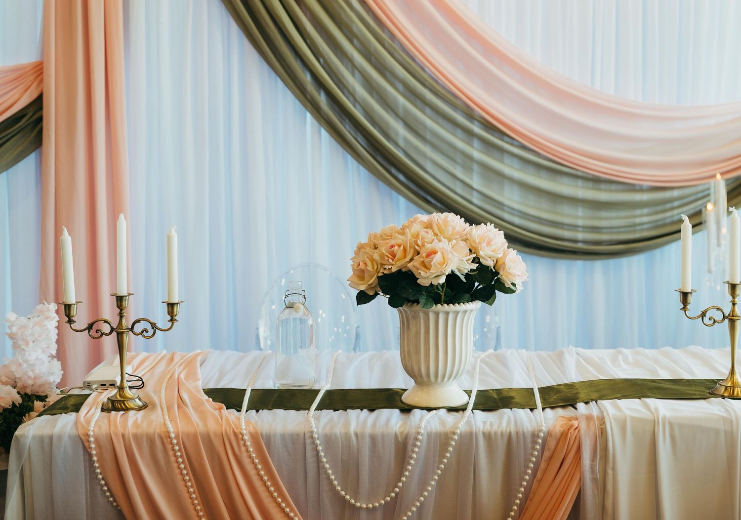 Elegant wedding table with peach roses, candelabras, pearl garland, and draped peach and gray fabric backdrop.