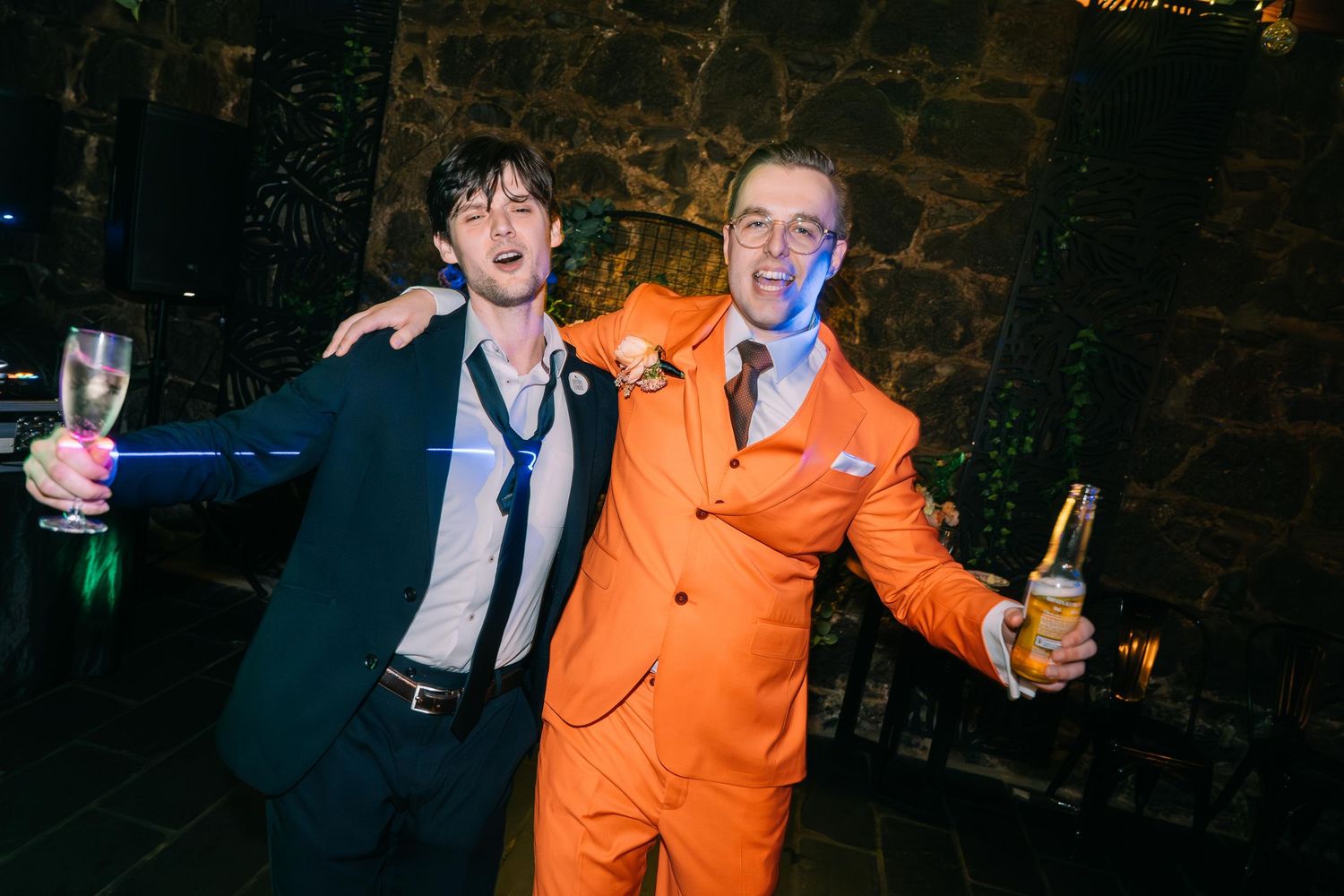 Two men smiling and posing at a wedding party, one wearing an orange suit holding a beer.