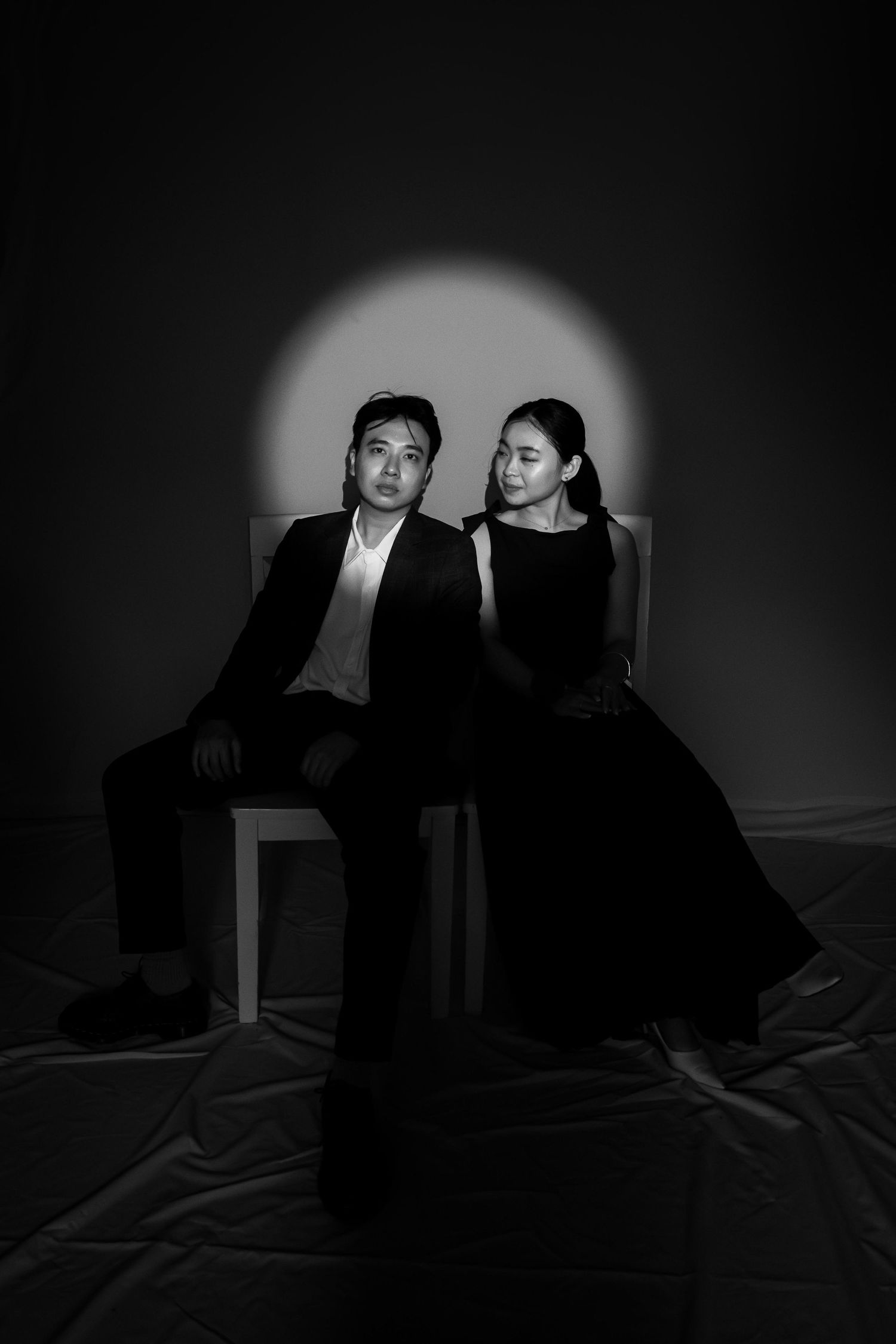 Black and white portrait of a man in a suit and woman in a black dress seated together under a spotlight.