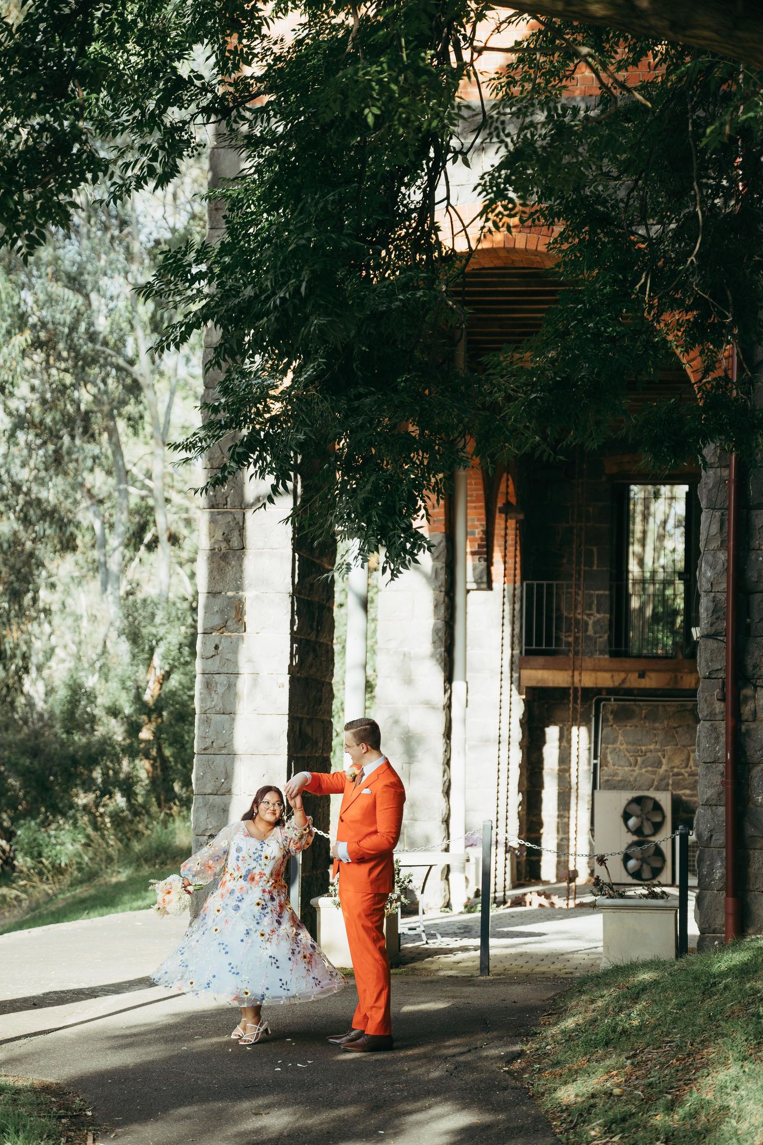 Bride in floral dress and groom in orange suit dance playfully outside a stone building among trees.