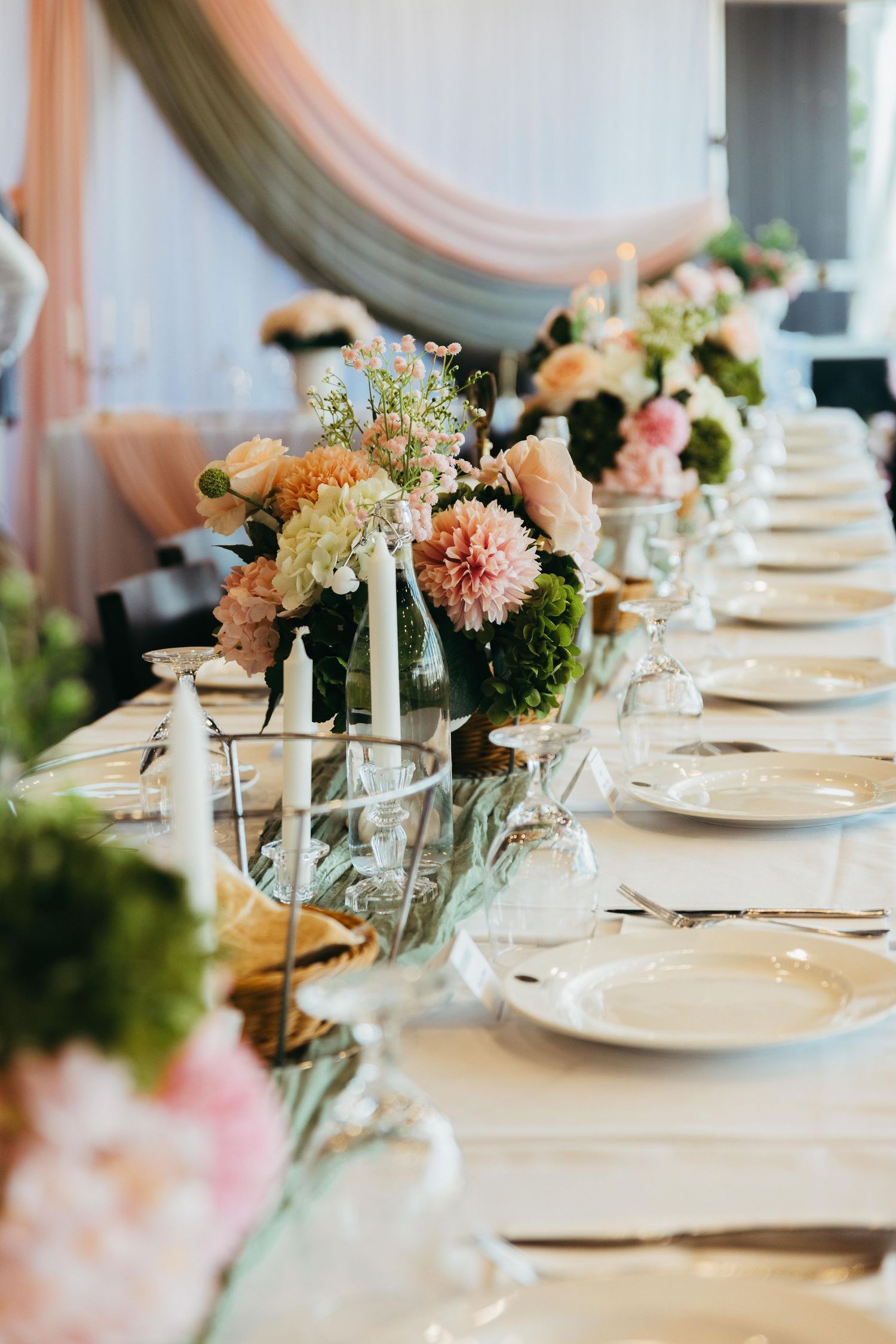 Romantic wedding banquet table with white plates, candles, and lush pink and green floral arrangements under draped fabric.