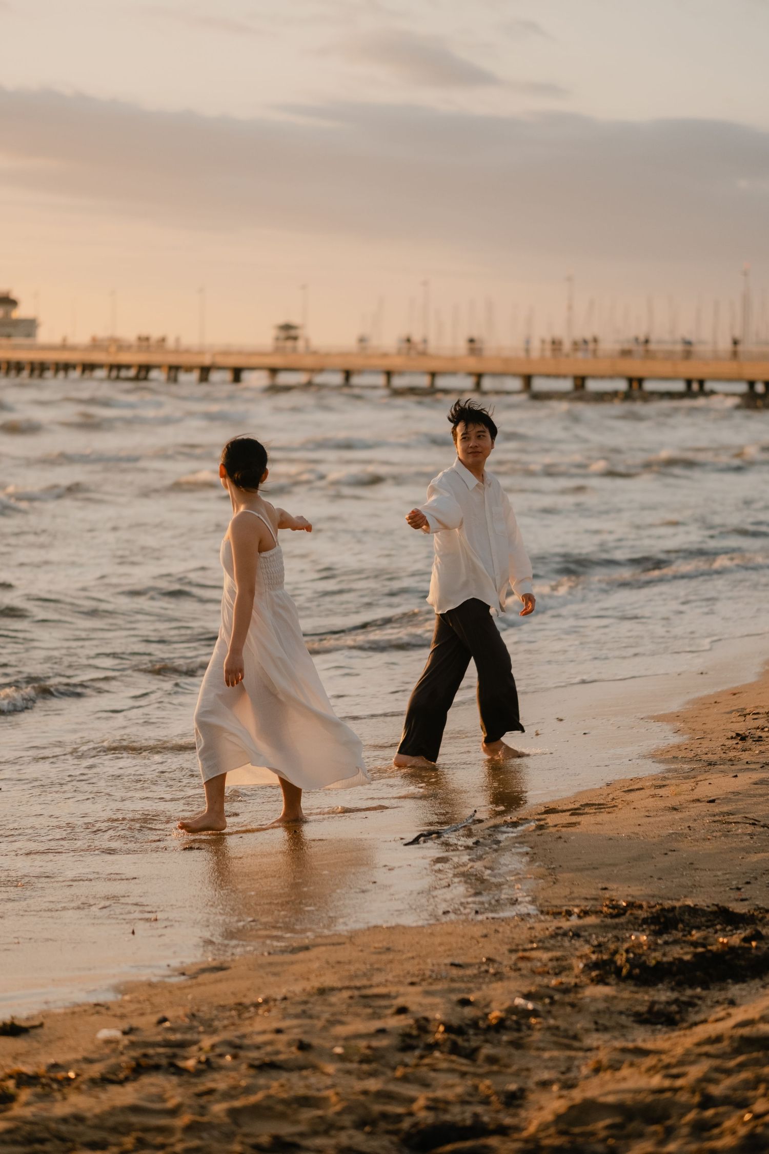Couple playing in ocean waves at sunset on sandy beach, woman in flowing white dress, man in white shirt.