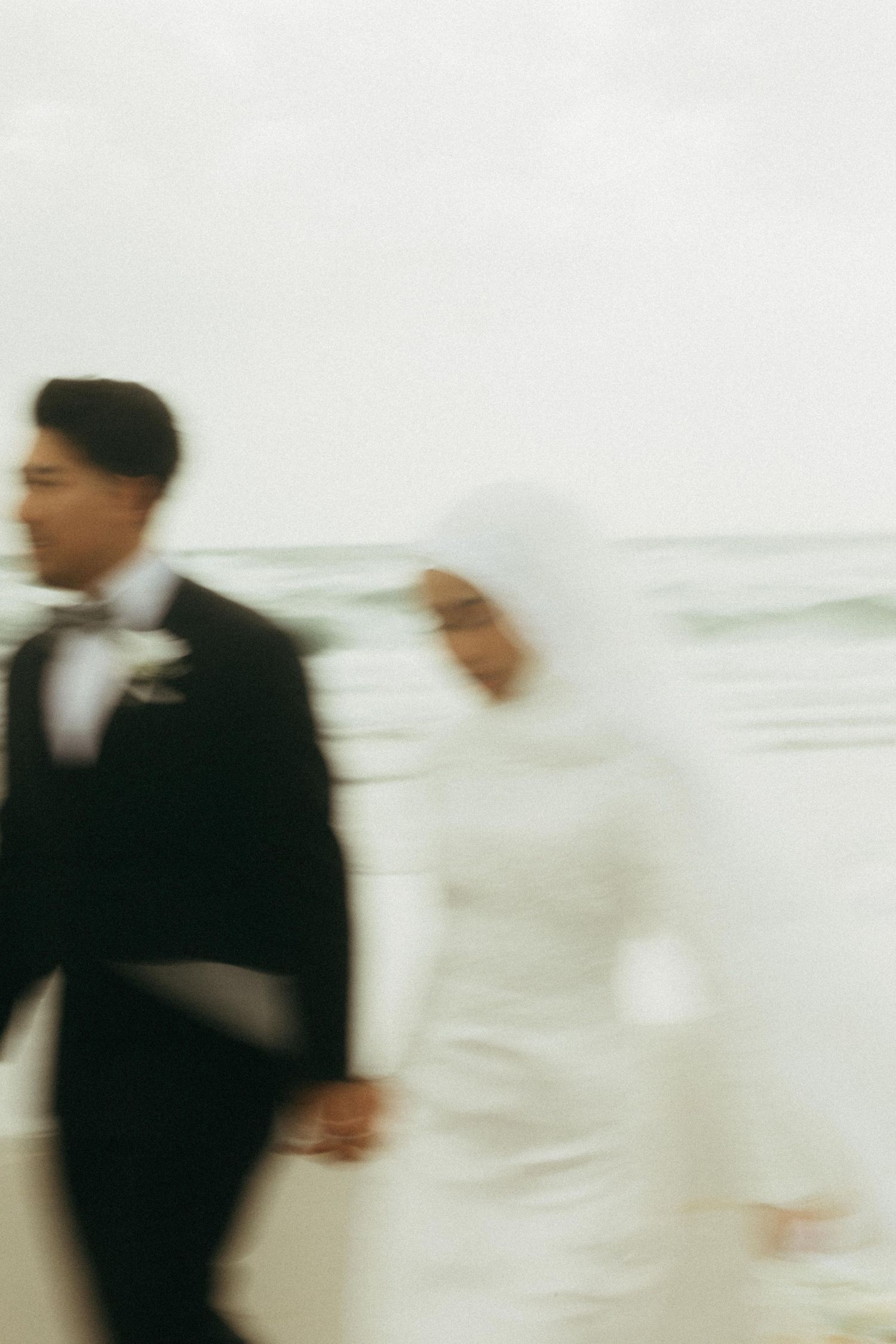 Blurred motion photo of Muslim bride and groom holding hands walking along beach.