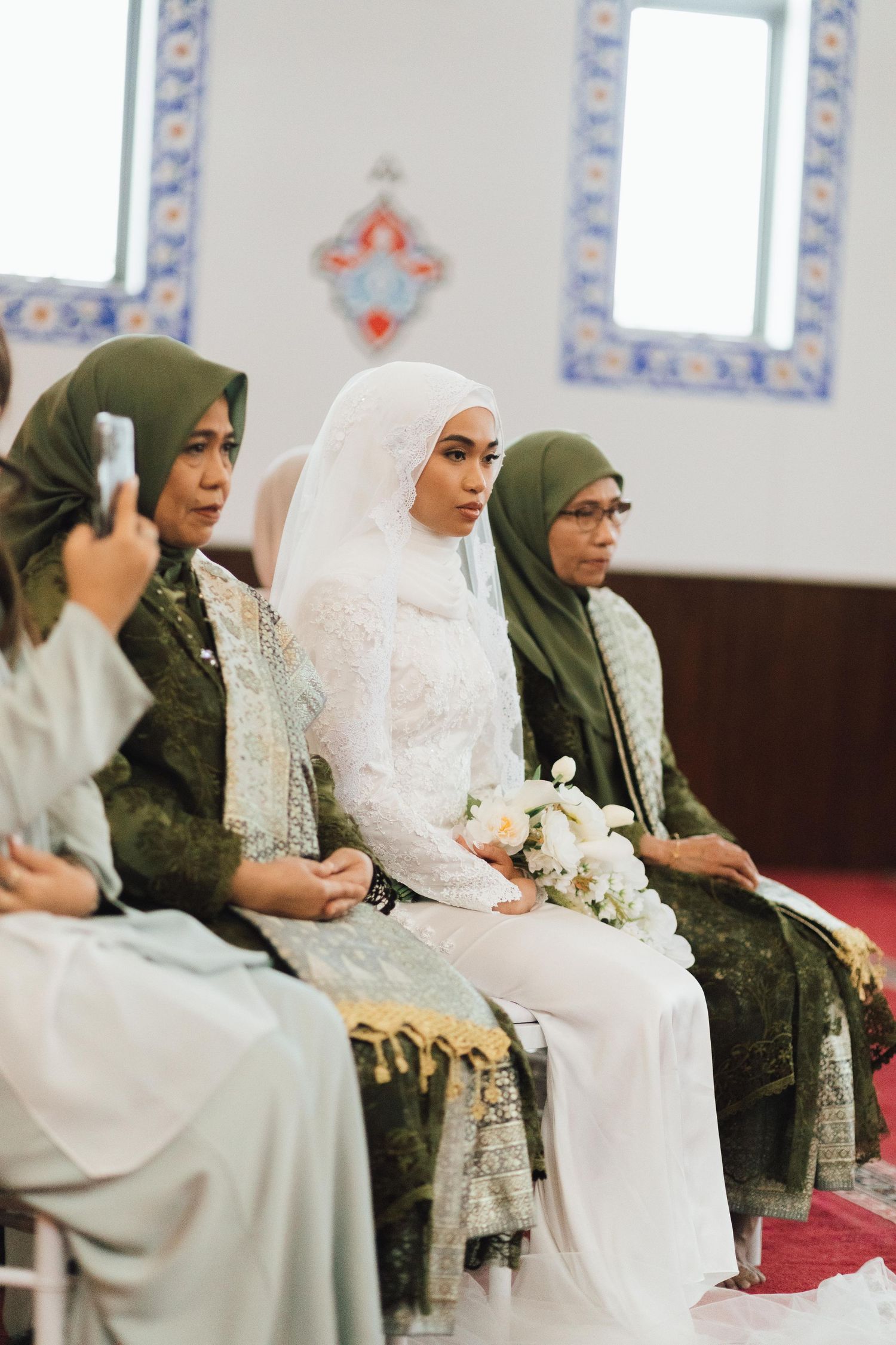 A Muslim bride in white hijab and gown holds flowers while seated with women in green hijabs at an Islamic wedding ceremony.