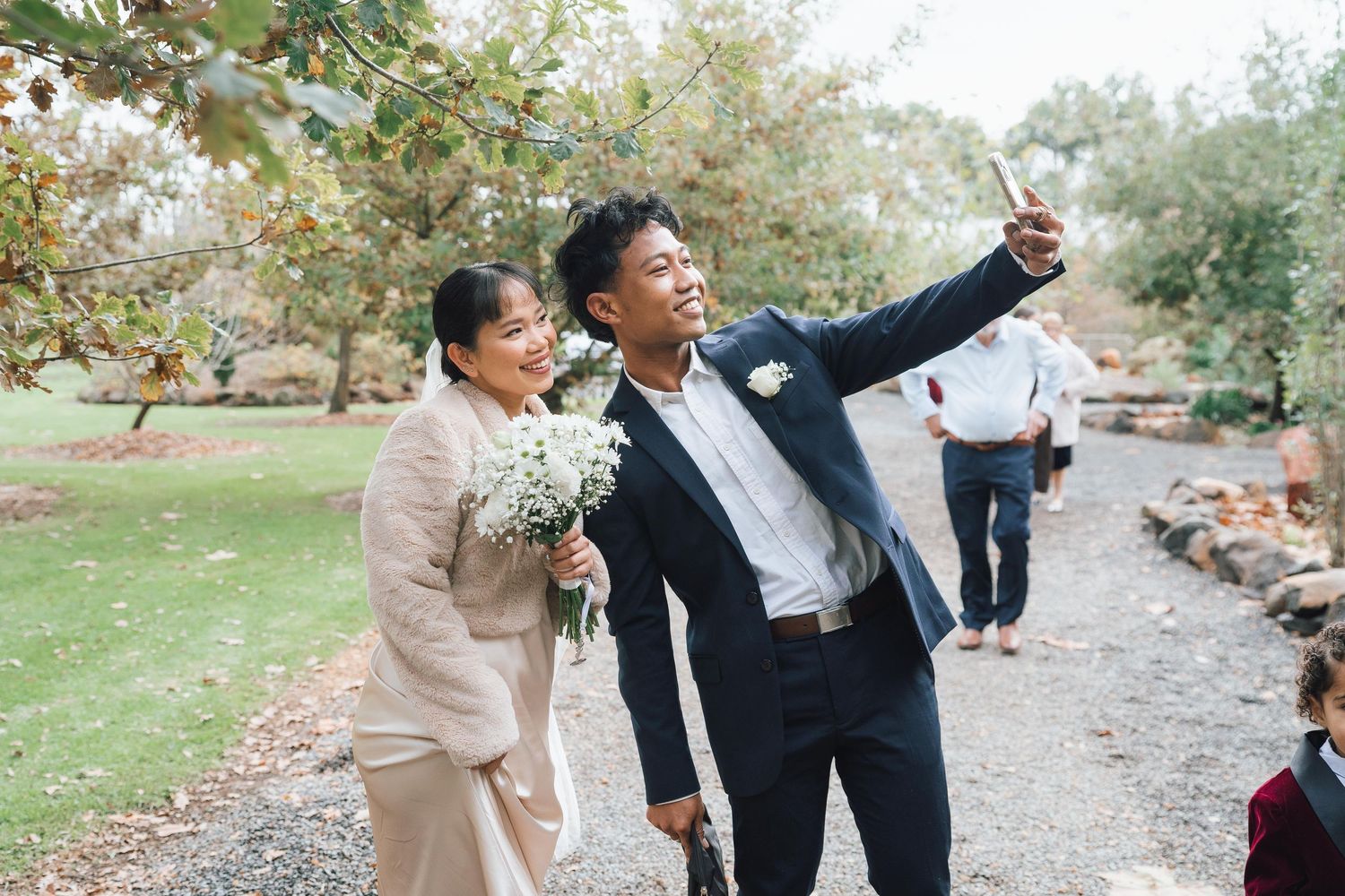 Groom in navy suit takes selfie with woman holding white bouquet at outdoor wedding venue.