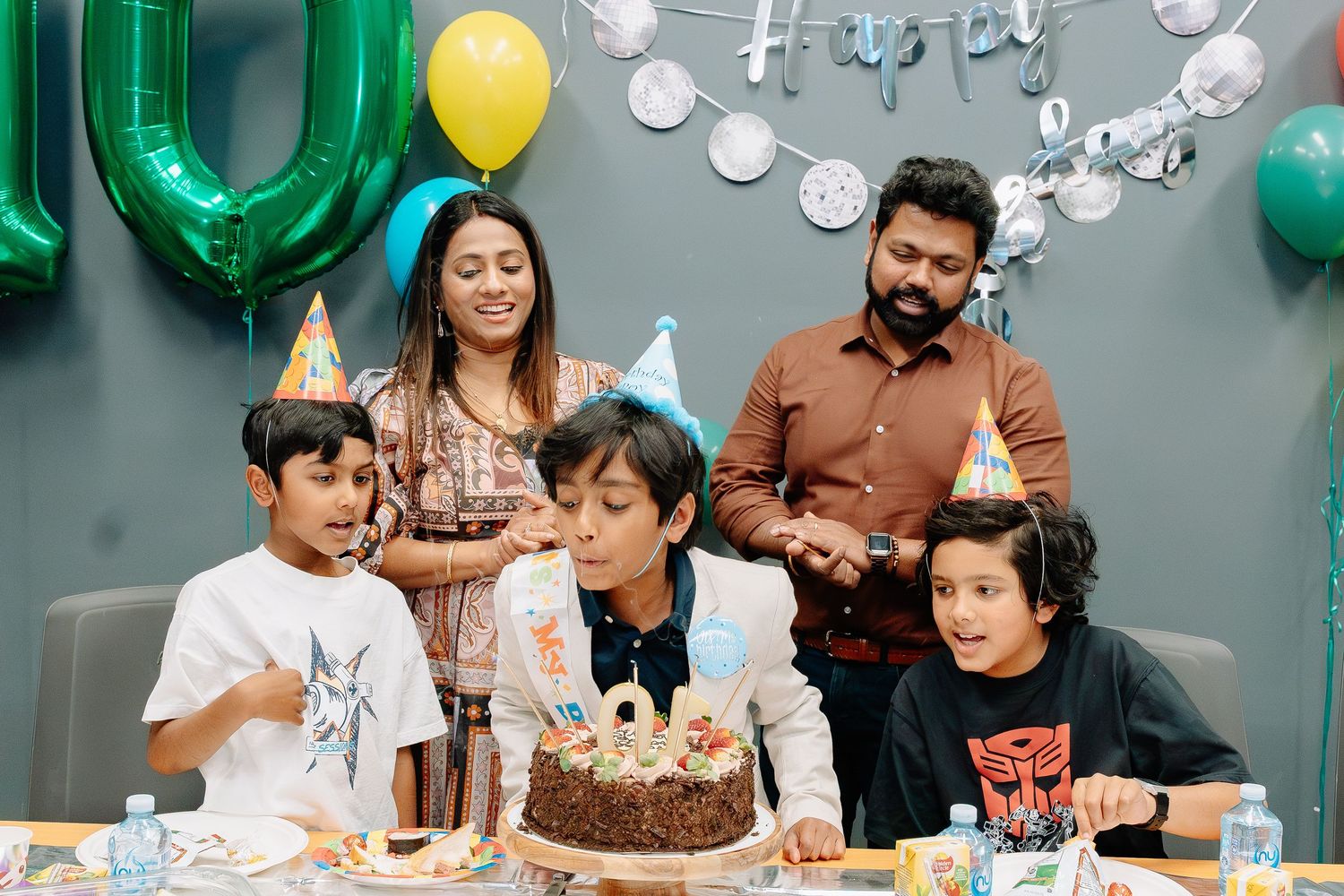 Family celebrates special occasion with chocolate birthday cake surrounded by green and yellow balloons and decorations.