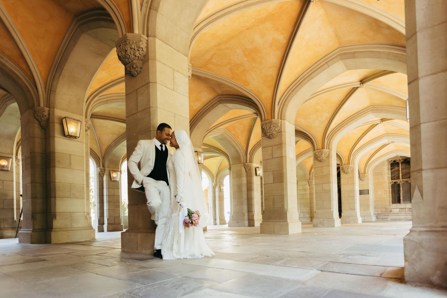 Bride and groom pose under elegant stone arched colonnade in a grand historic building during wedding photography session.