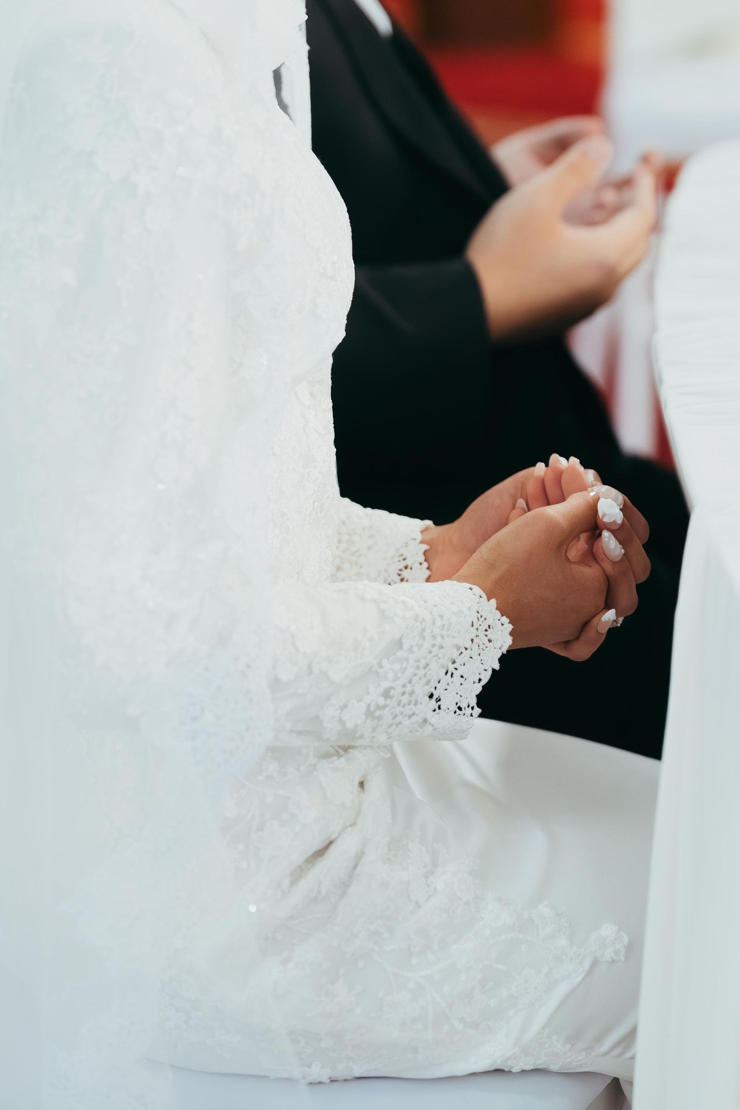 A bride in white lace hijab gown clasps hands during a Muslim wedding ceremony.