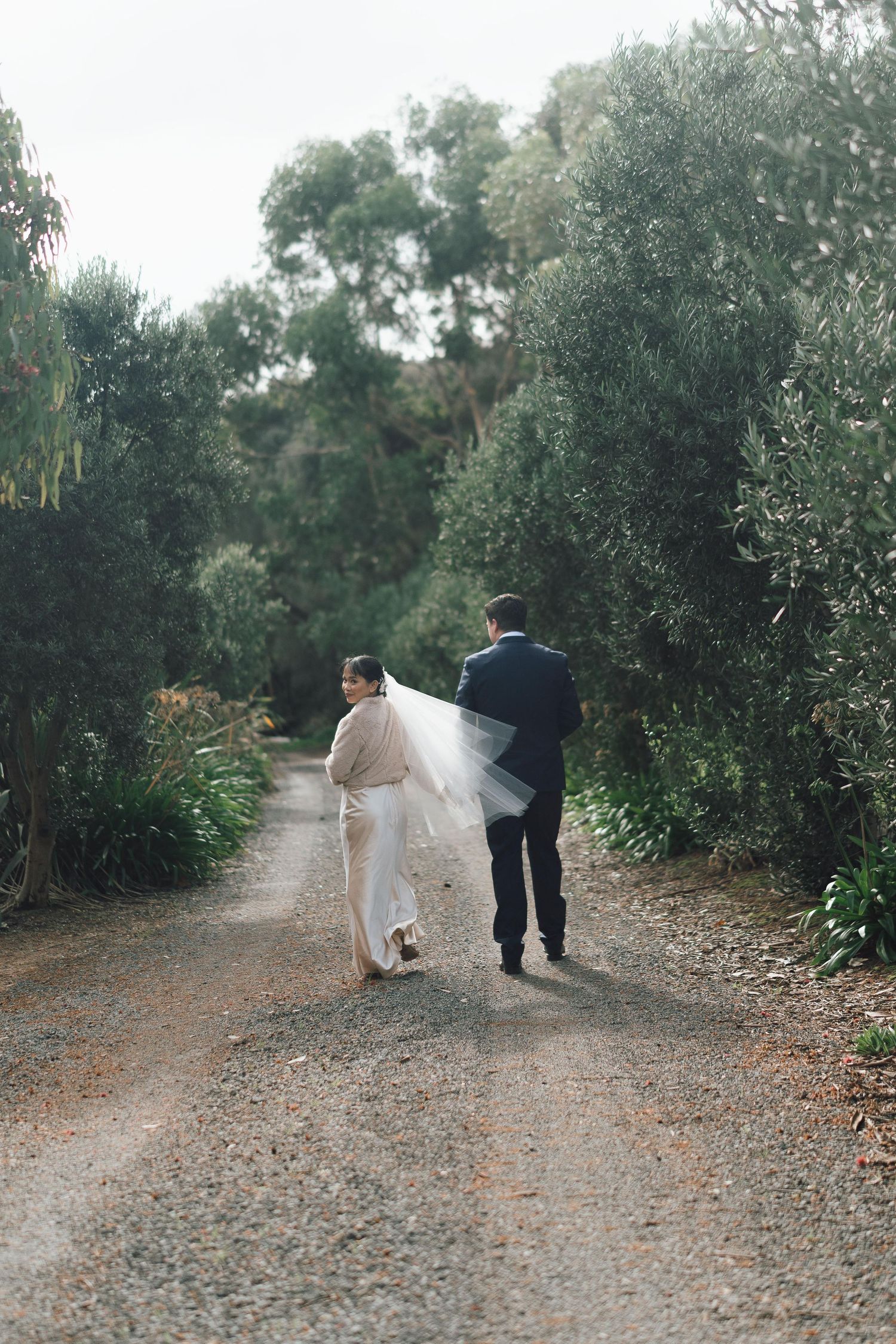 Bride and groom walk hand in hand down a tree-lined gravel path after their outdoor wedding ceremony.