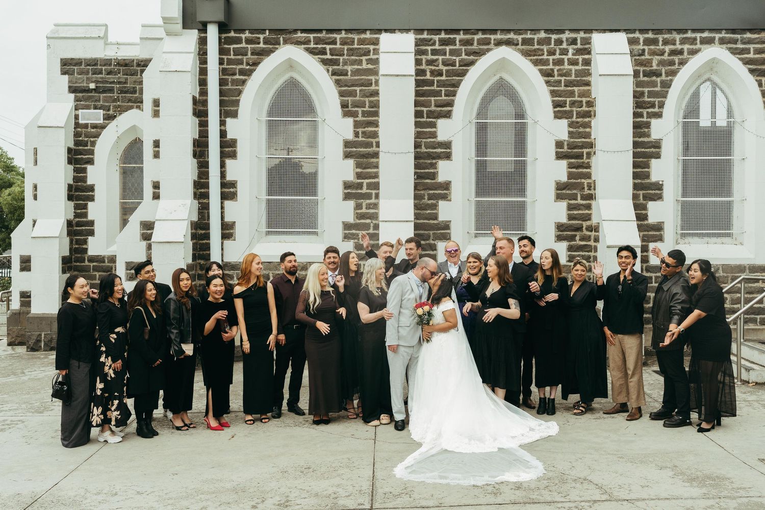 Bride and groom kiss surrounded by wedding party in black attire outside a historic stone church.
