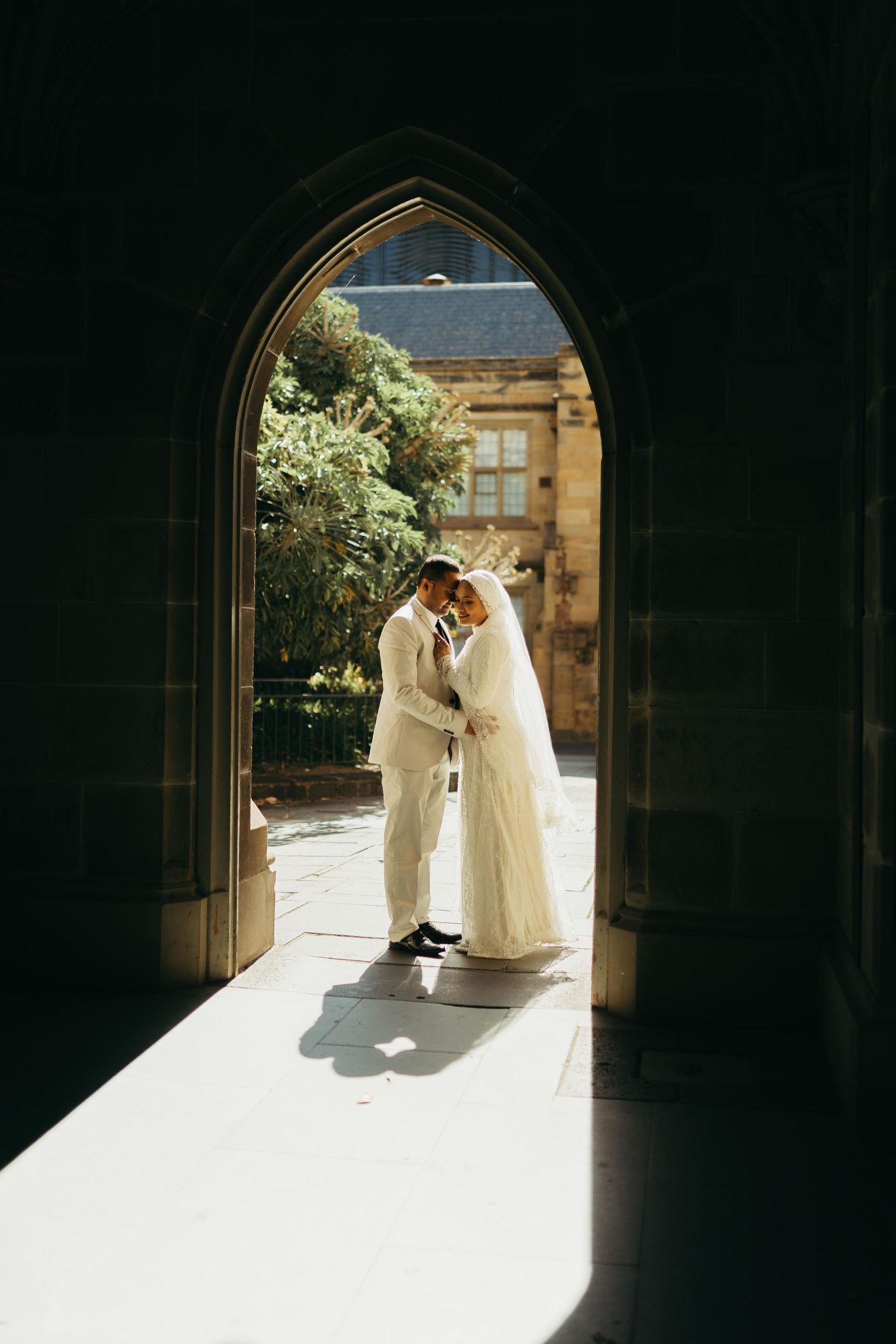 Bride and groom sharing a romantic moment framed by a Gothic stone archway in sunlight.