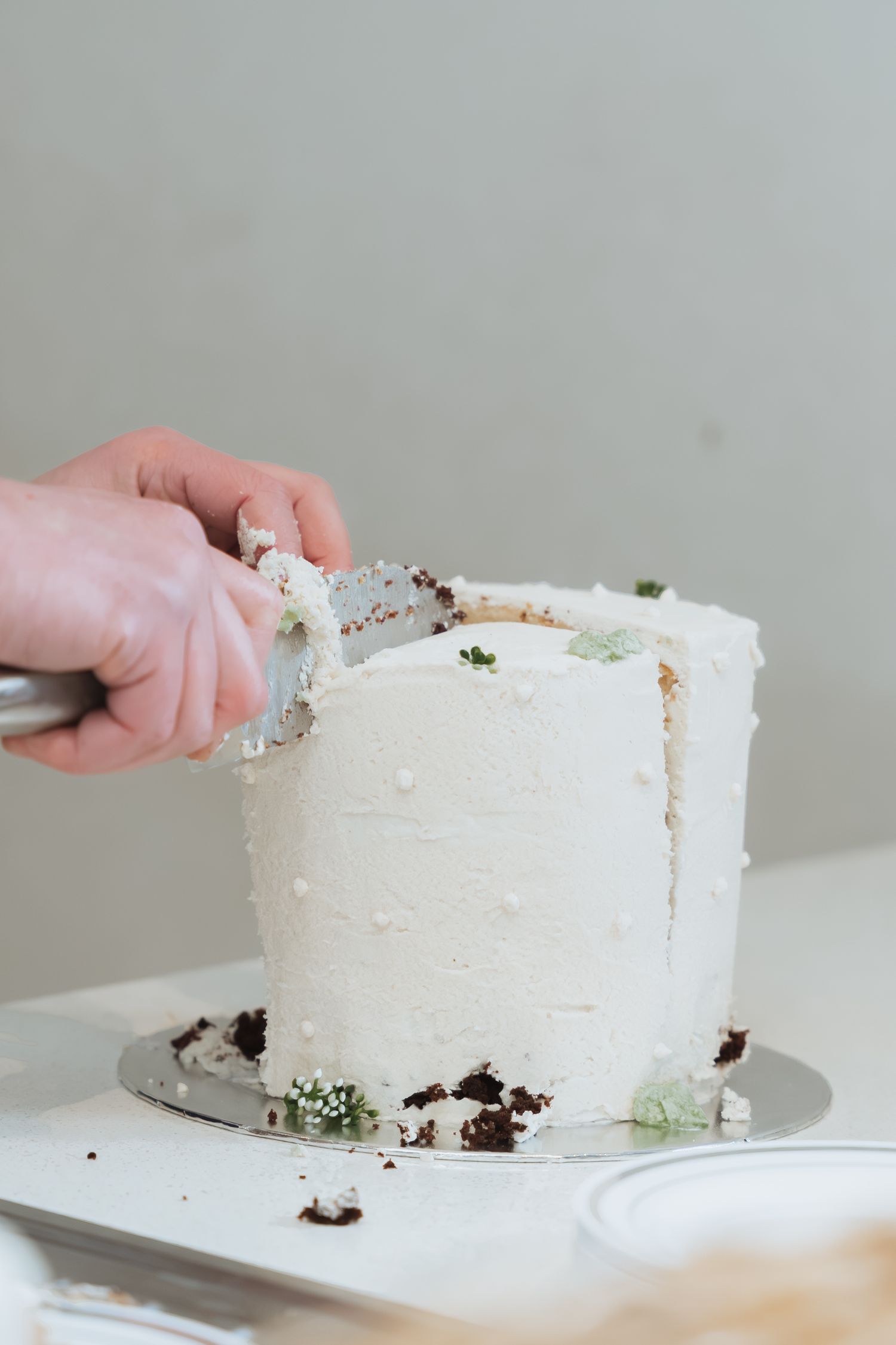 A hand using an offset spatula to frost a white layer cake on a cake stand.