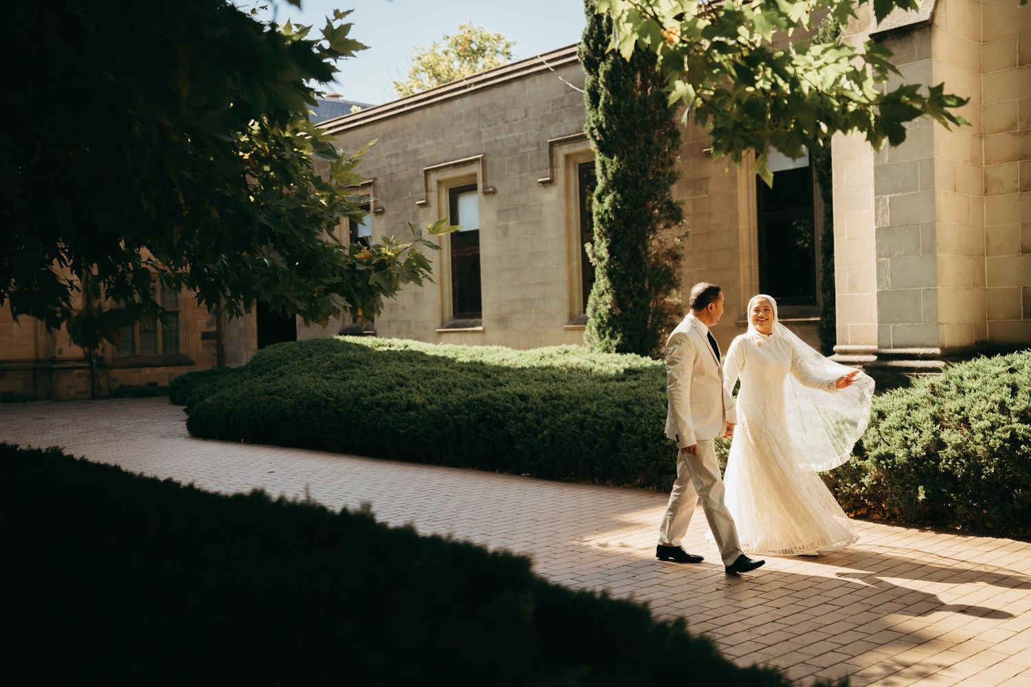 Bride in flowing white gown and groom walk together along sunlit garden pathway.