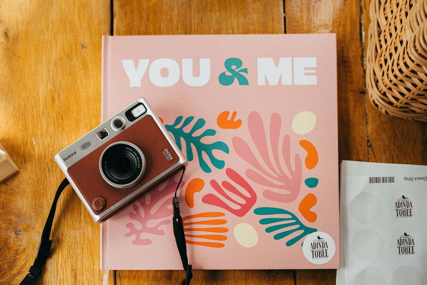 Instax mini camera on a pink 'You & Me' photo book with colorful floral designs on a wooden surface.