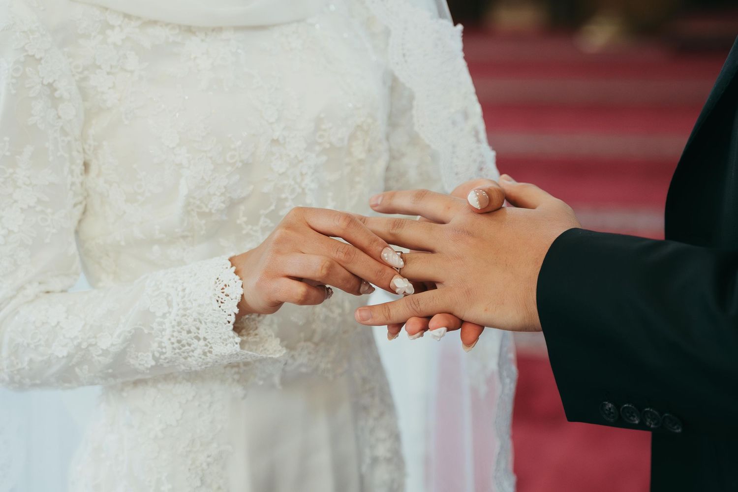 Bride in white lace dress placing wedding ring on groom's finger during marriage ceremony.