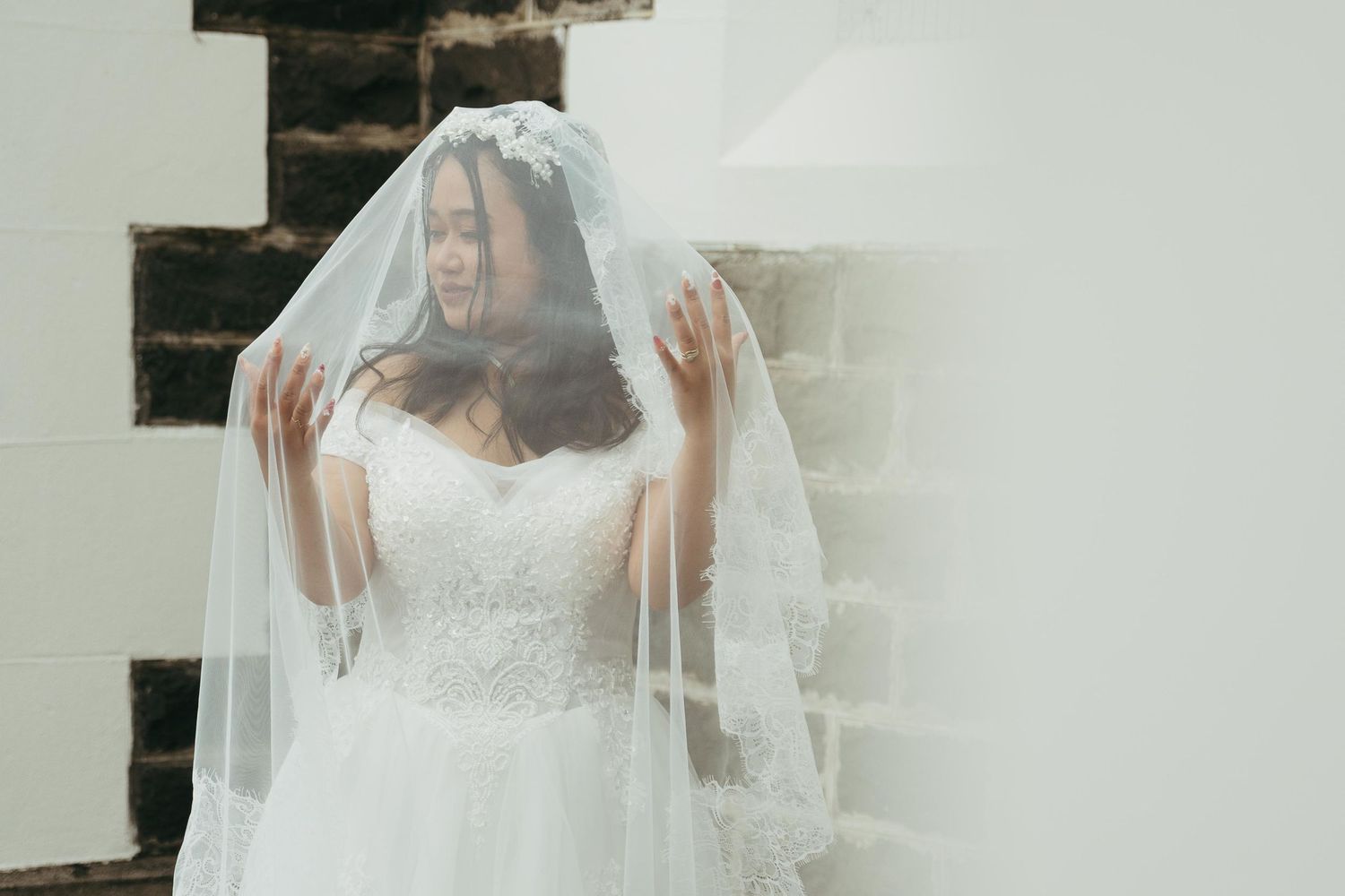 Bride in lace wedding dress and flowing veil posed against stone wall in elegant bridal portrait.