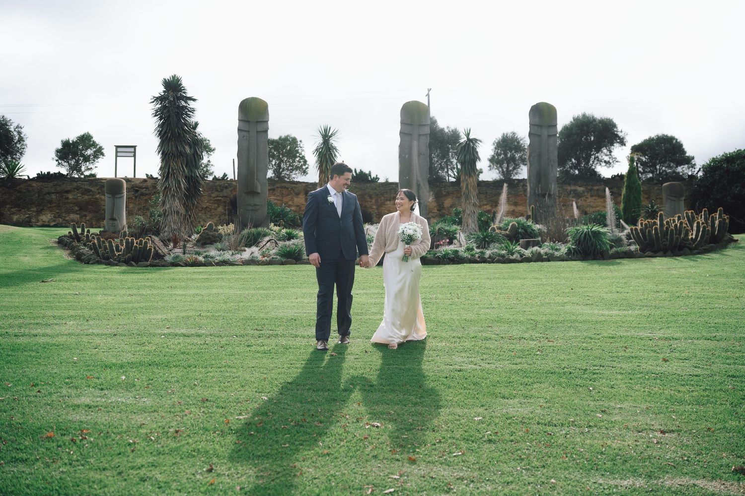 Bride and groom walk hand in hand on lush green lawn at scenic outdoor wedding venue.