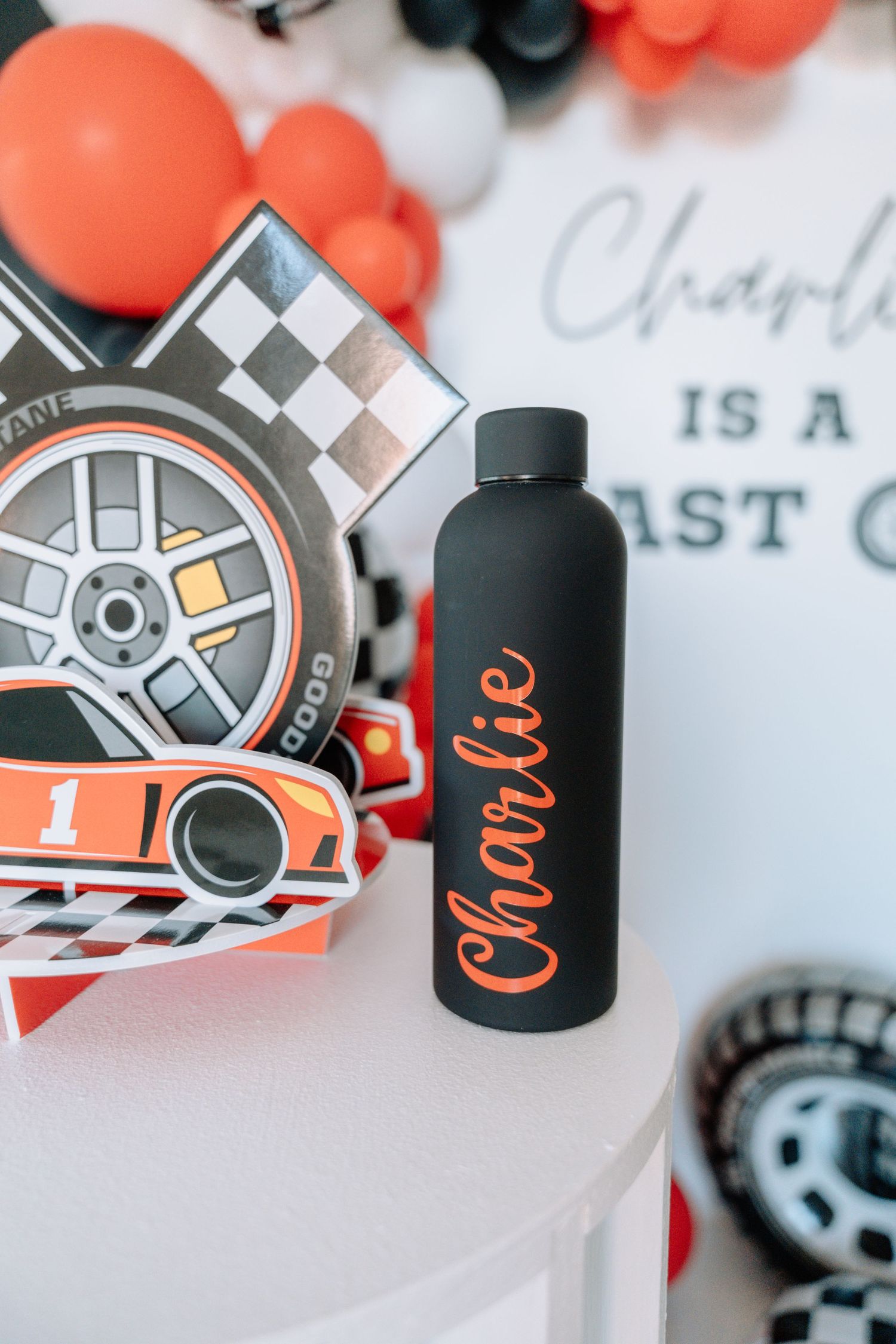 Black water bottle with orange text sits next to racing decorations and colorful balloons at a party.