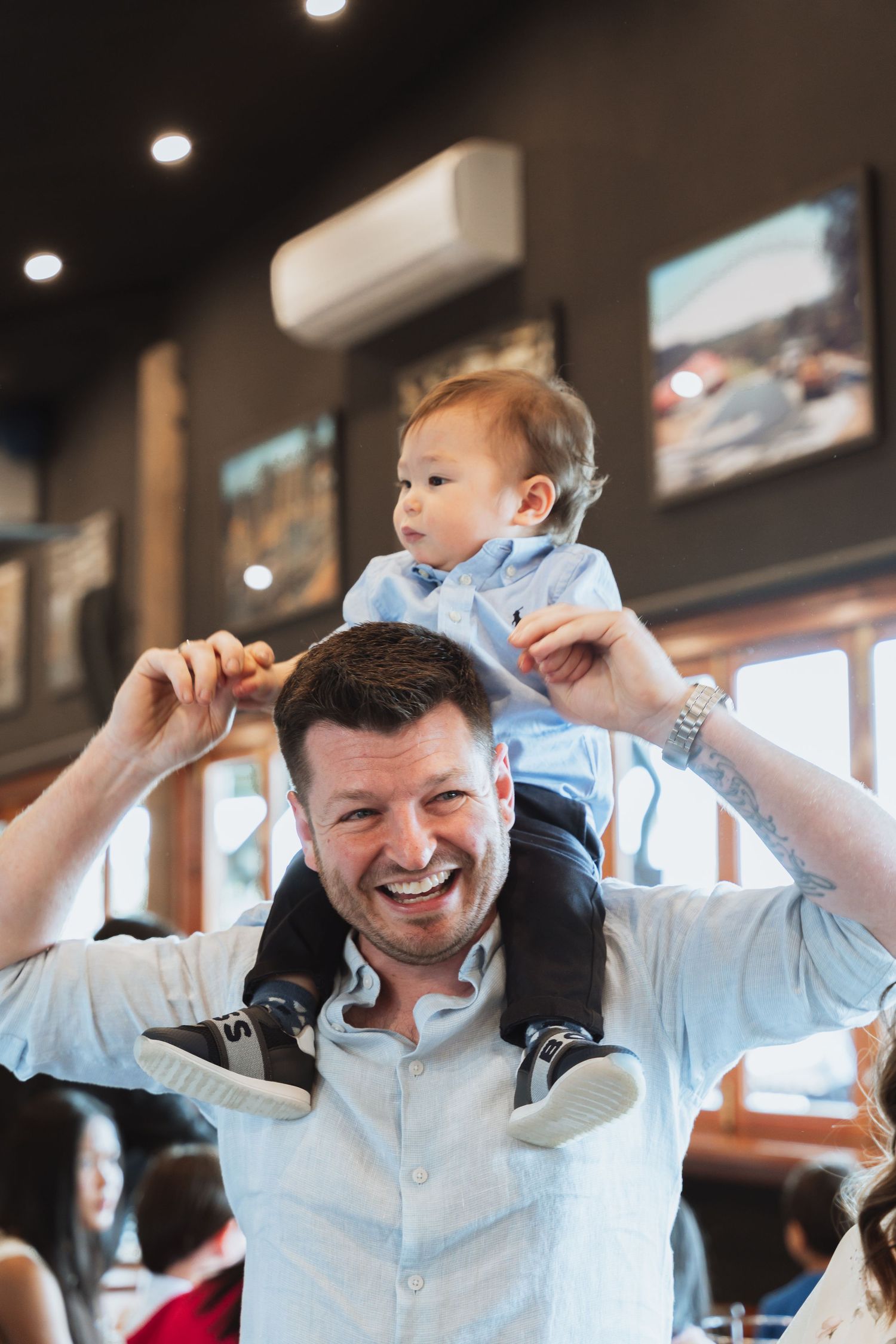 A joyful moment captured at a restaurant as a baby rides on shoulders while surrounded by overhead lighting.