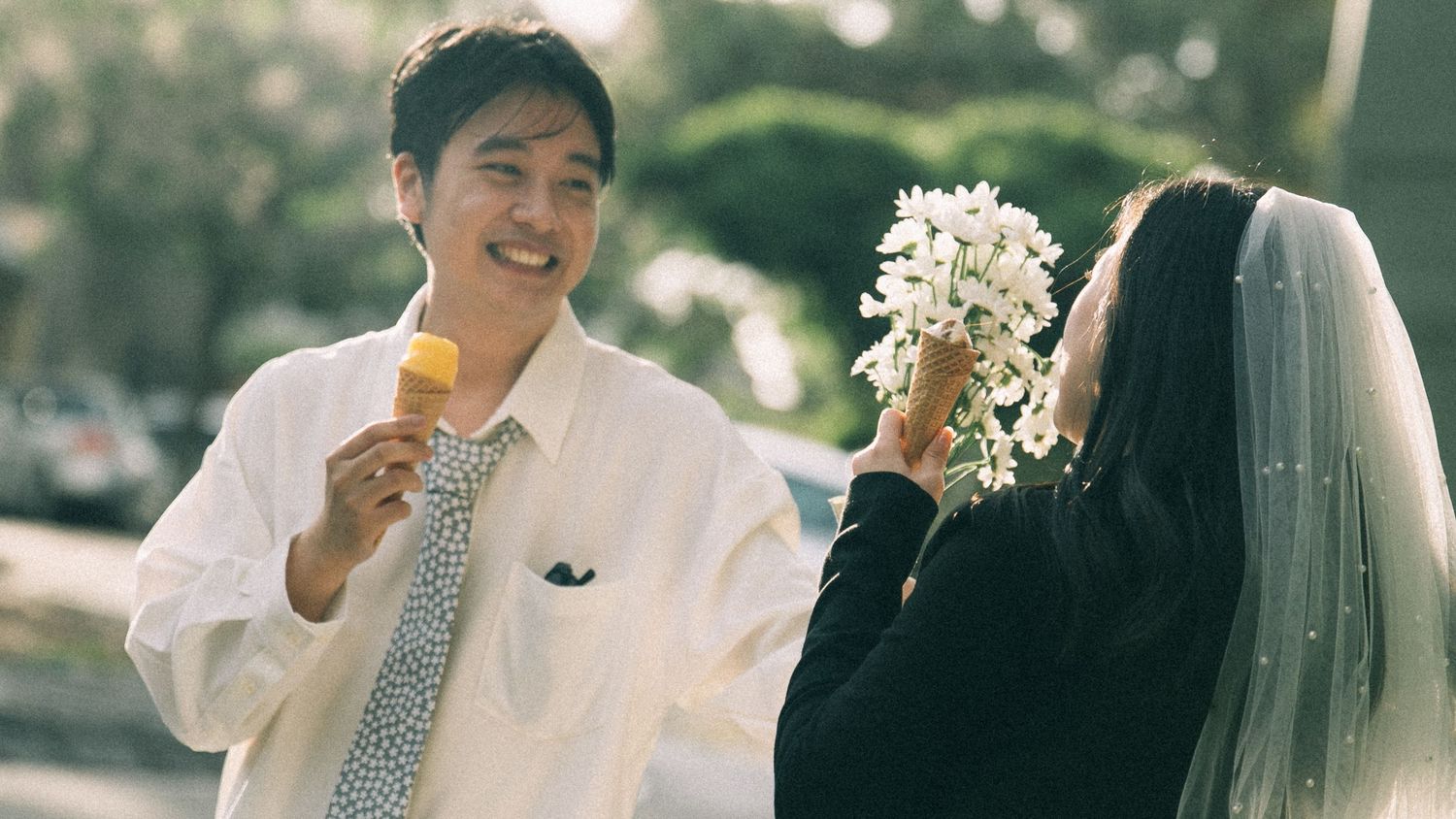 Groom holding ice cream smiles at bride with white flower bouquet and veil outdoors on sunny day.