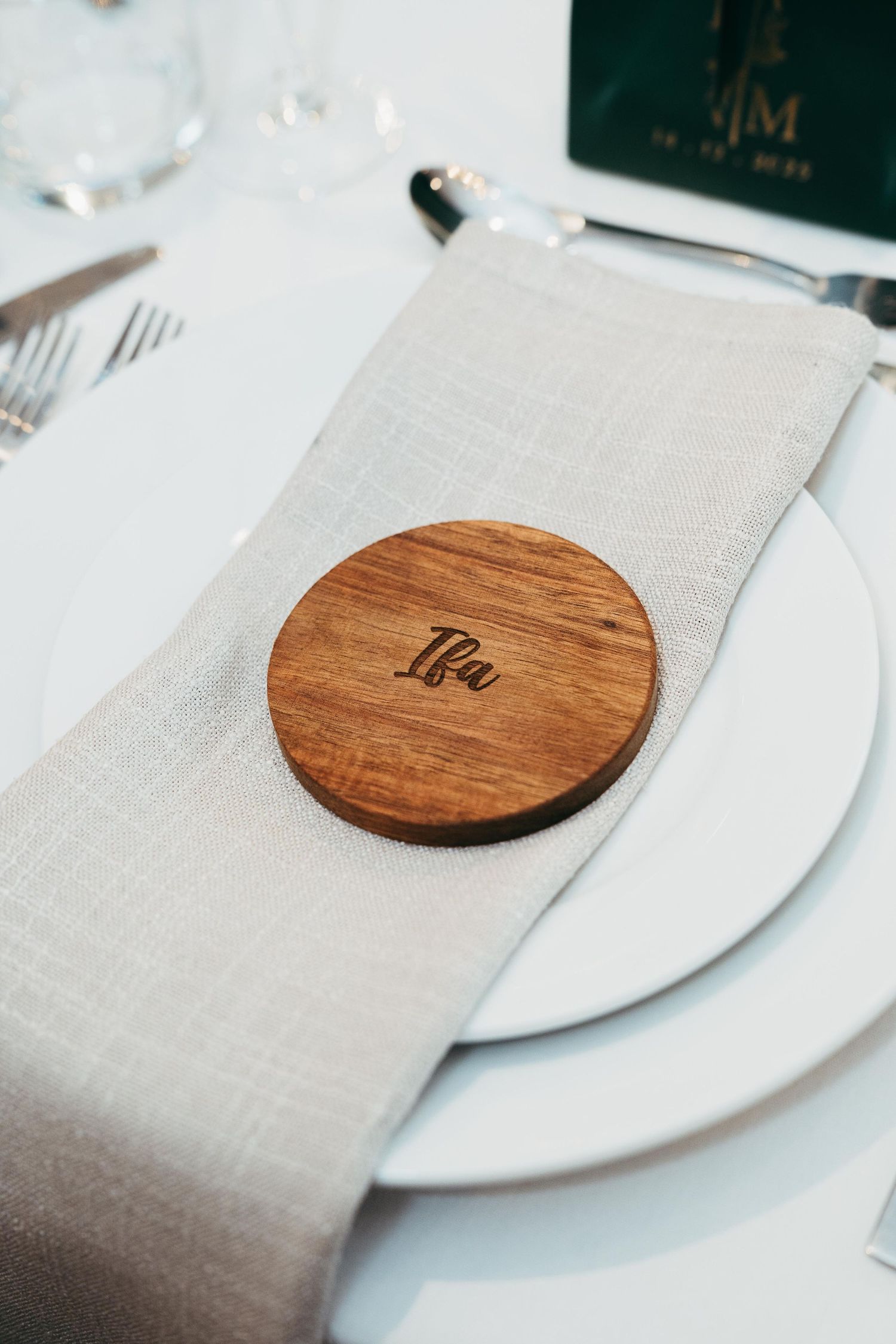 Elegant wedding place setting with engraved wooden coaster name card reading Ifa on a linen napkin and white plate.