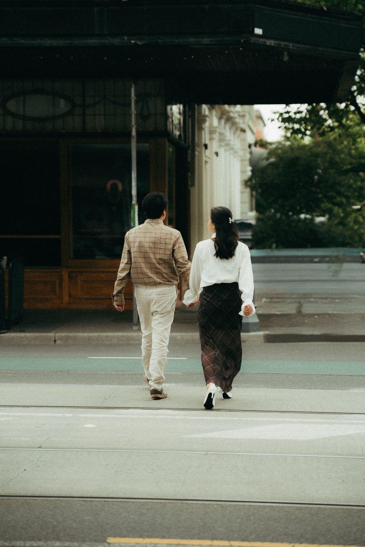 Couple holding hands crossing a city street, viewed from behind, wearing casual elegant attire.