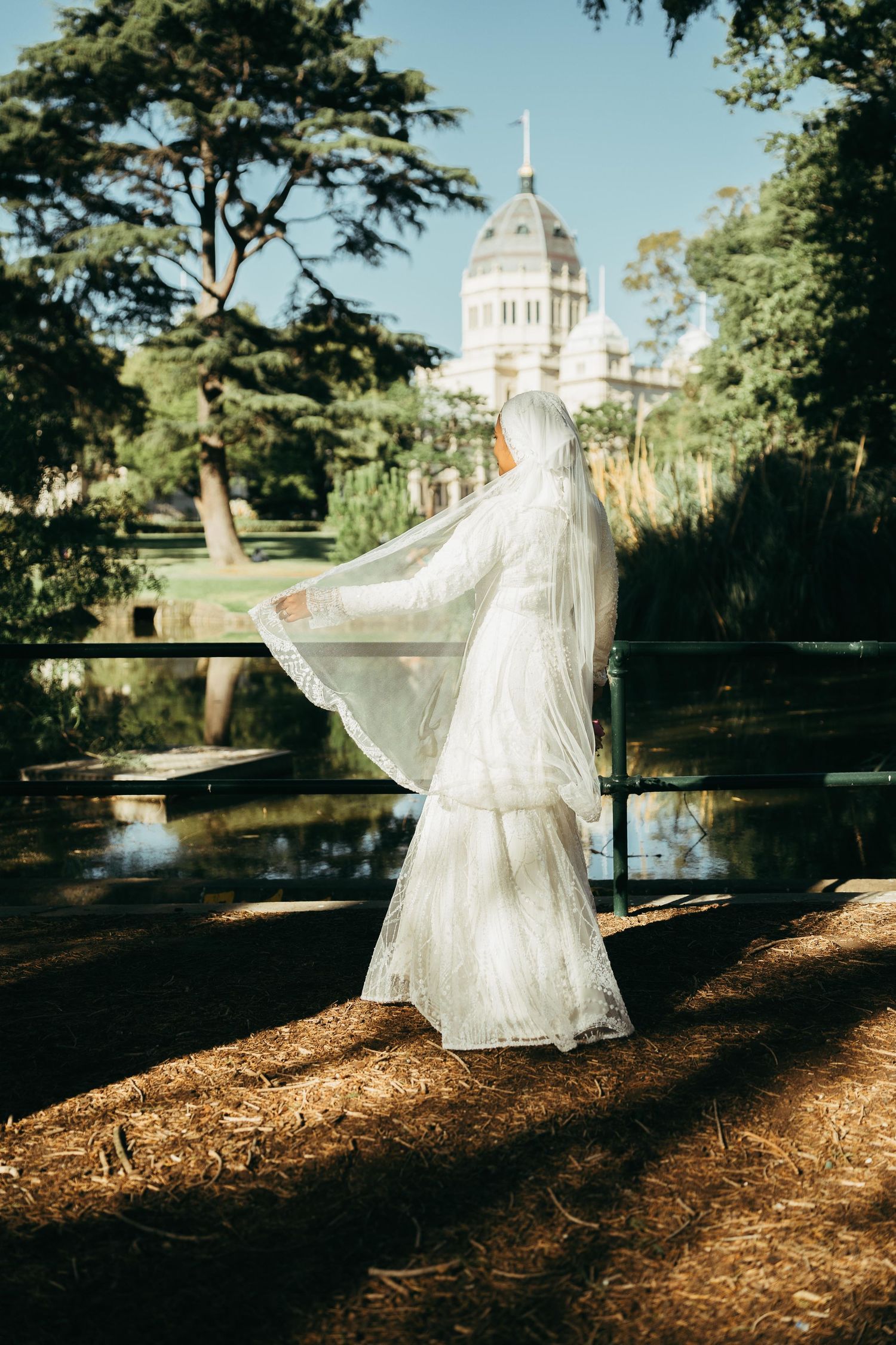Muslim bride in white lace gown and flowing veil standing by a pond with a domed building backdrop.