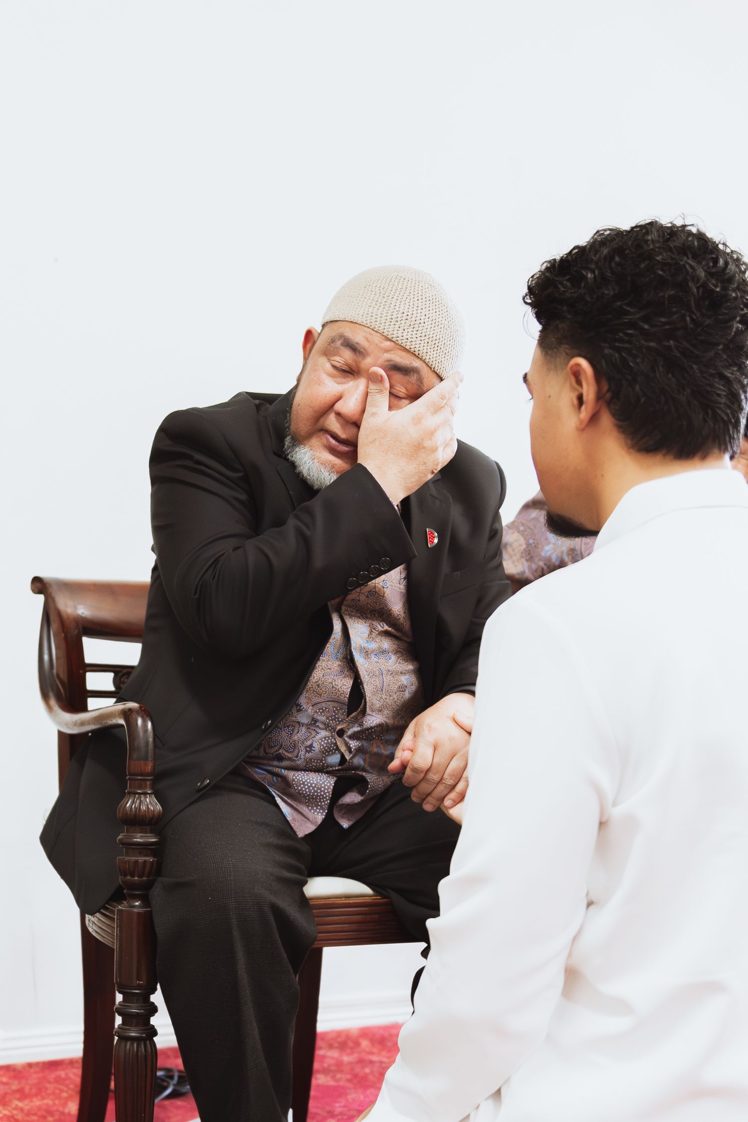 Elderly man in black suit and kufi cap wiping eye while speaking with younger man in white shirt.