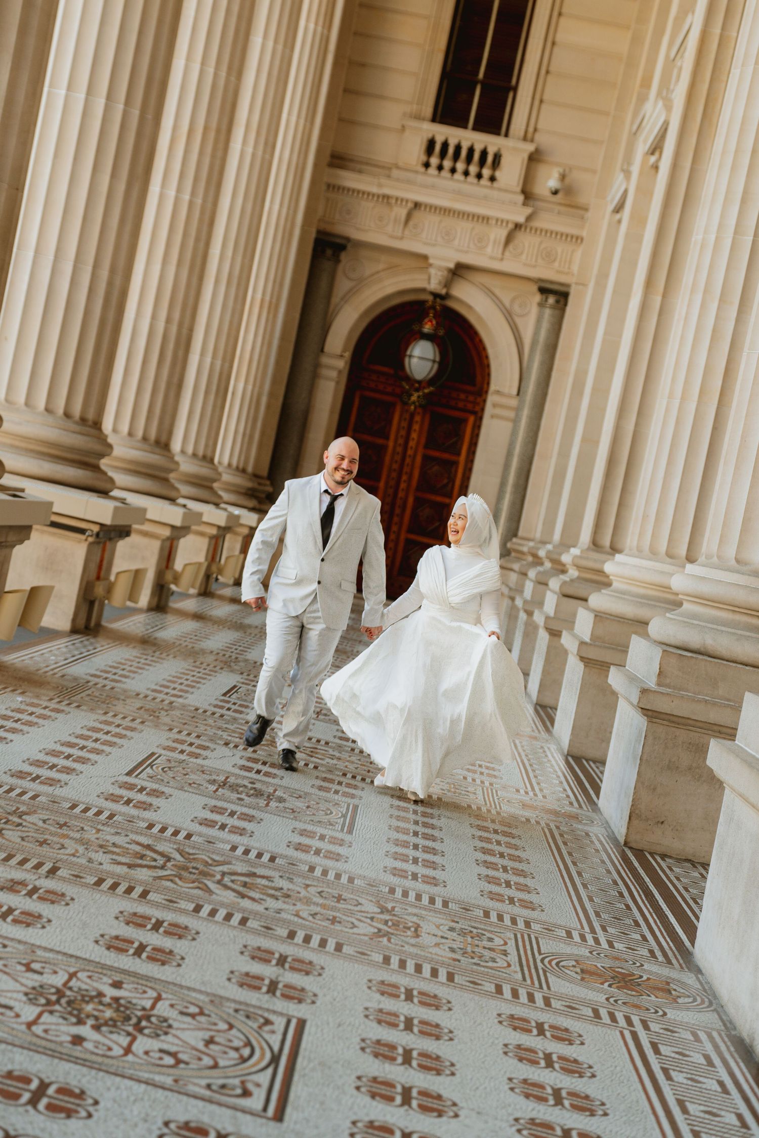A Muslim bride in white hijab and groom in light suit walk joyfully on ornate tiled steps beside grand columns.