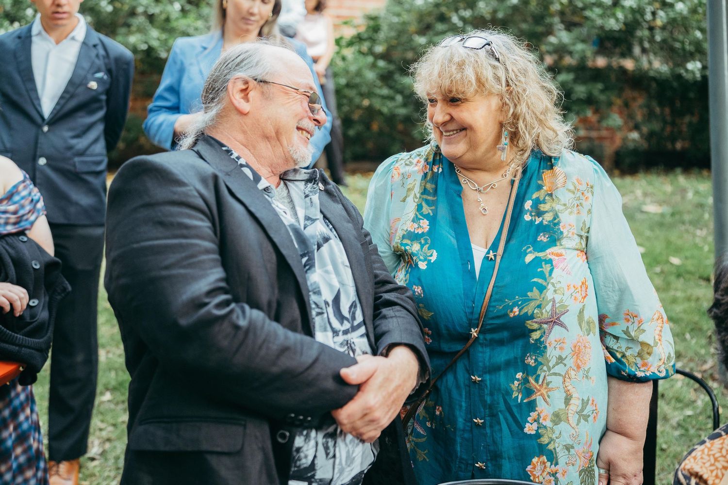 Two elderly wedding guests laughing and holding hands warmly in a sunlit garden.
