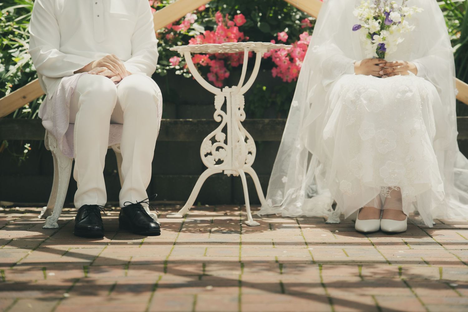 Bride and groom sitting on a bench in a garden, wearing wedding attire with white shoes and black dress shoes.